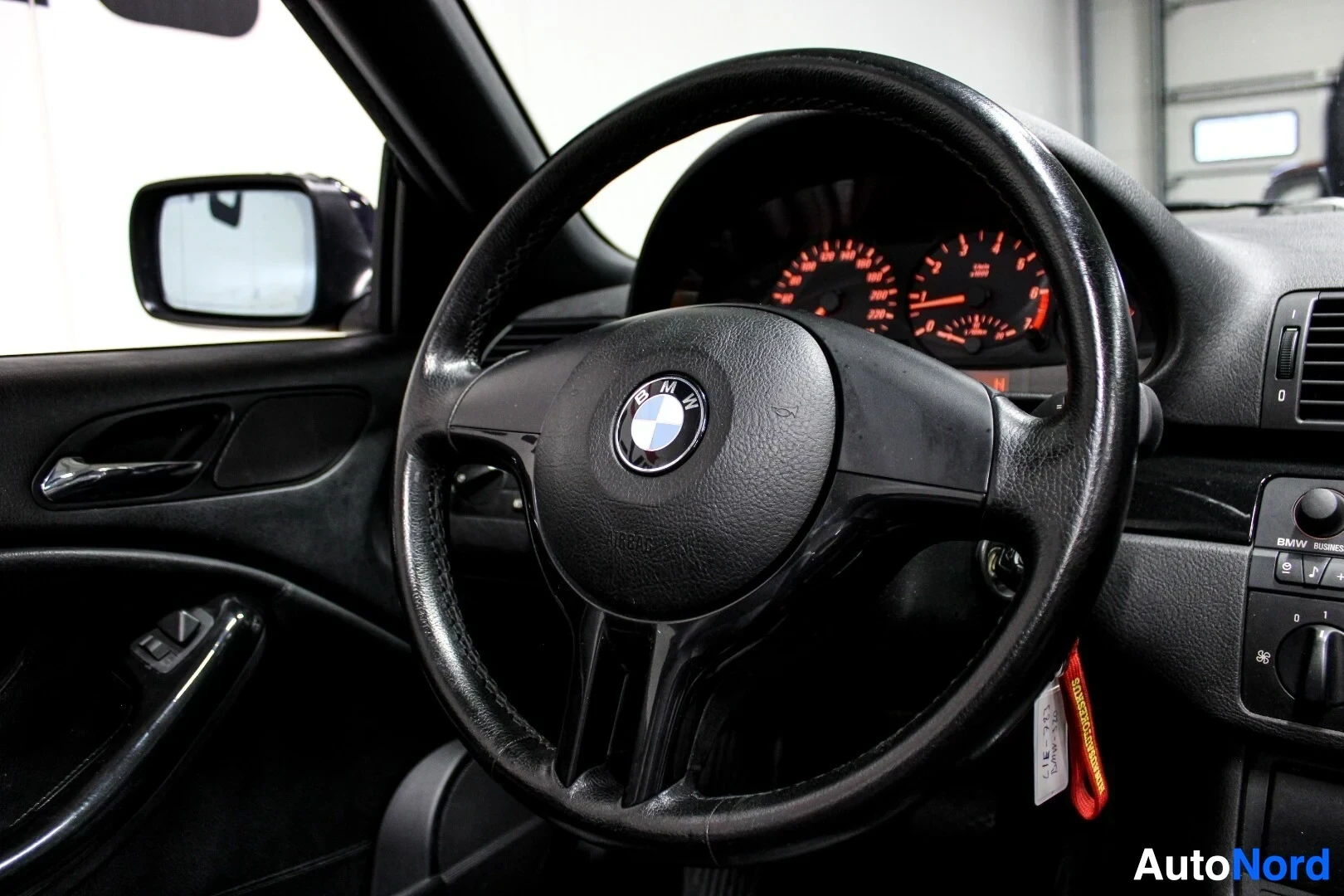 Yksityiskohta: Lähikuva auton mustasta ohjauspyörästä, jonka keskellä on BMW-logo, sekä mittaristosta, jossa on oranssi taustavalo. Peilin heijastus näkyy vasemmalla.