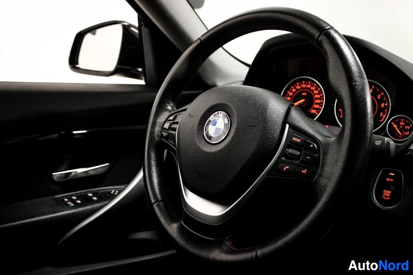 Yksityiskohta: Monitoimiohjauspyörä vasemmalta edestäpäin, mittaristo taustalla, ohjauspyörän painikkeet, BMW-logo ja oven säätimet näkyvissä.