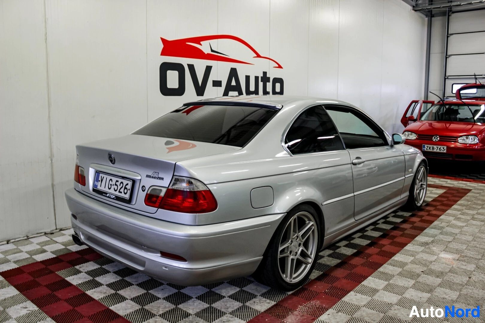 Takaviisto: Hopeanvärinen BMW, jossa on tummennetut takalasit ja AC Schnitzer -merkki takaluukussa, on pysäköity sisätiloihin valkoisen OV-Auto-logon eteen. Ajoneuvon rekis