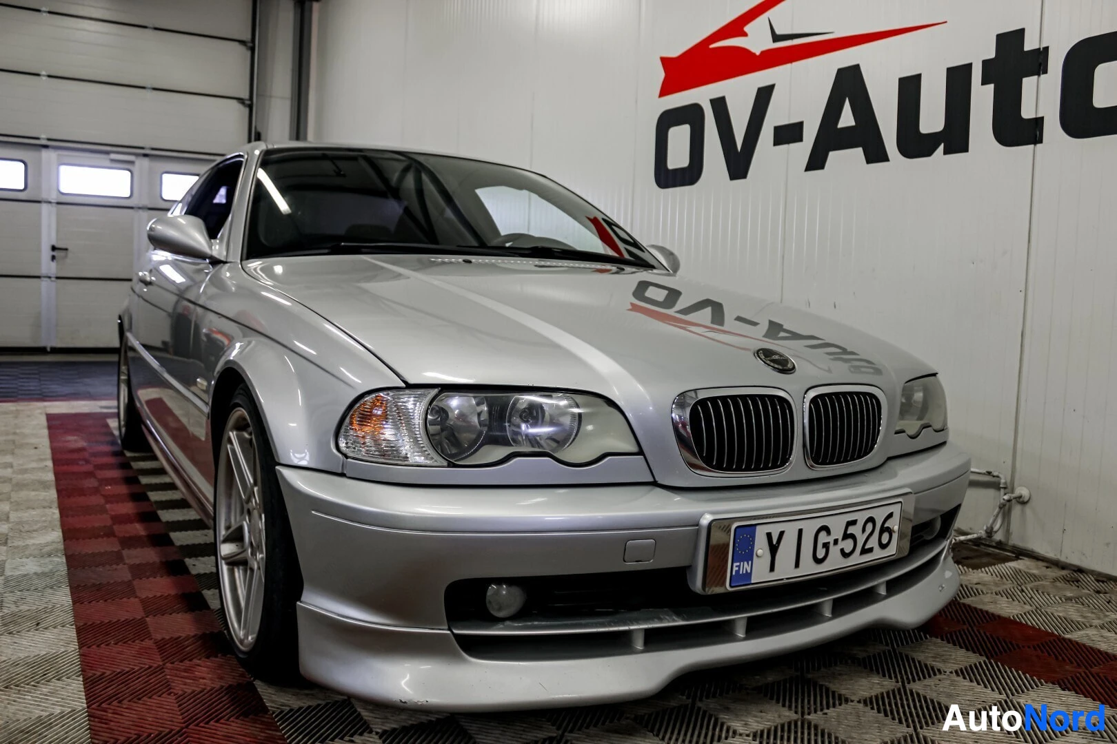Etuviisto, nokka vasemmalle: Hopeanvärinen BMW, jossa on suomalainen rekisterikilpi, on pysäköity sisätiloihin. Autossa on sporttinen etupuskuri ja kevytmetallivanteet. Tau