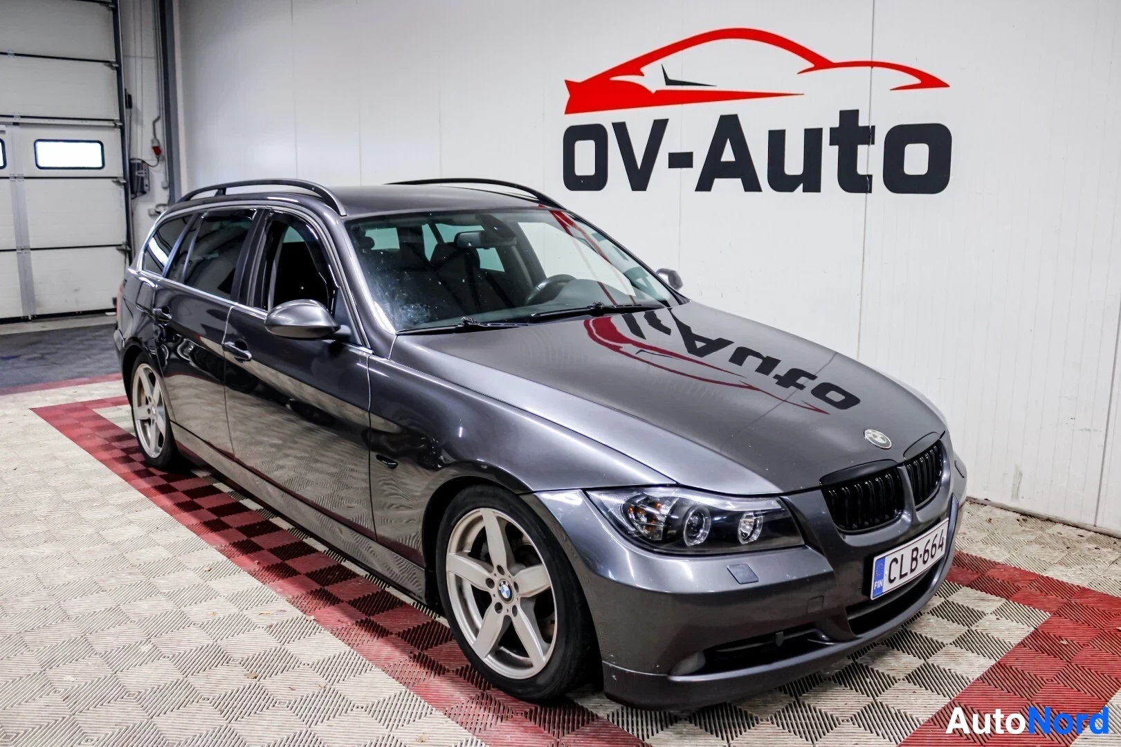 Etuviisto, nokka oikealle: Harmaa BMW 3-sarjan Touring pysäköitynä valkoisen ja punaisen kuvioidun lattian päälle autohalliin, jossa valkoinen seinä OV-Auto-logolla.
