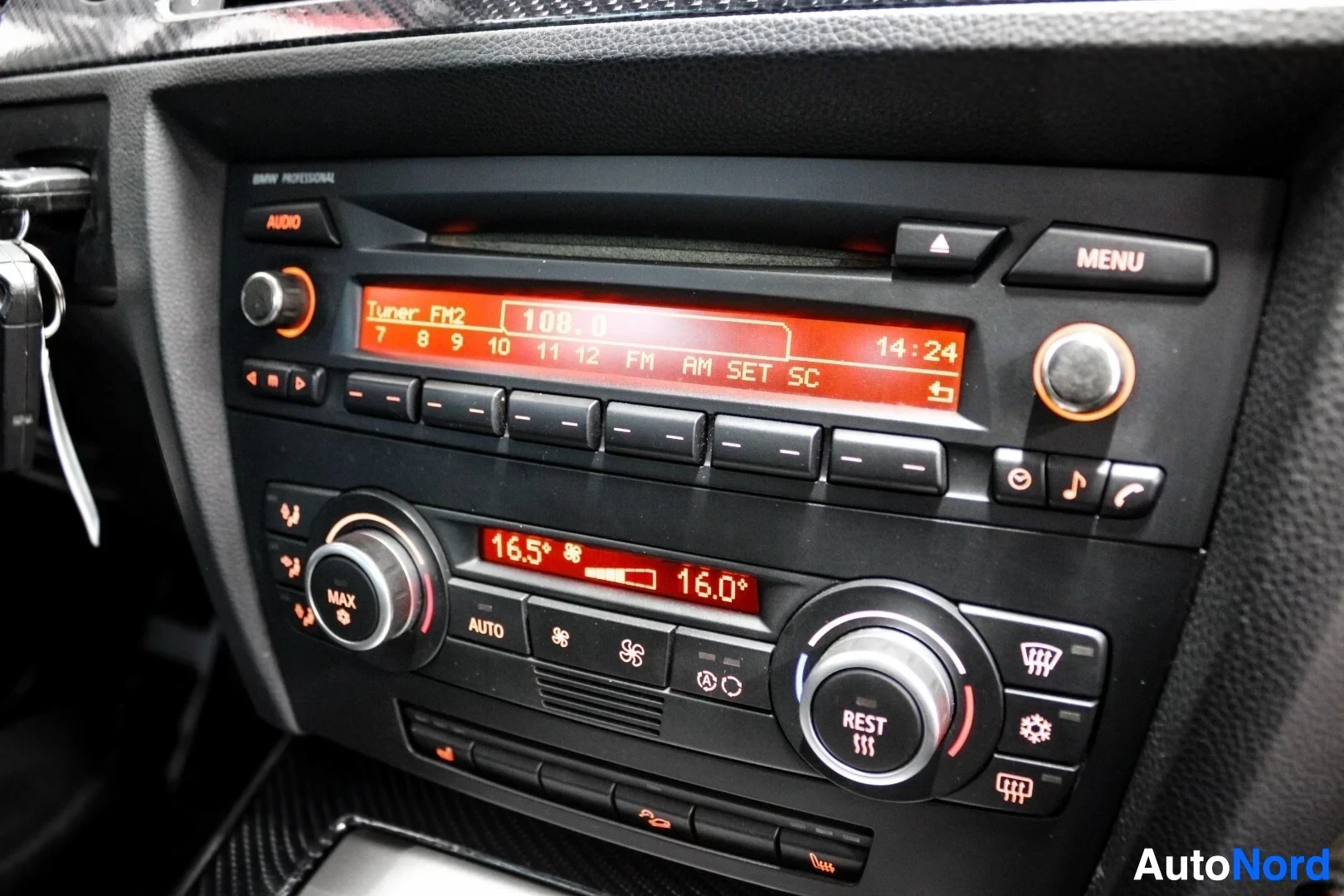 Ohjaamo: Lähikuva auton ohjaamon keskikonsolista, jossa näkyvät radio, joka näyttää taajuuden 100.8 ja ajan 14:24, sekä ilmastointilaitteen hallintalaitteet, joissa näky