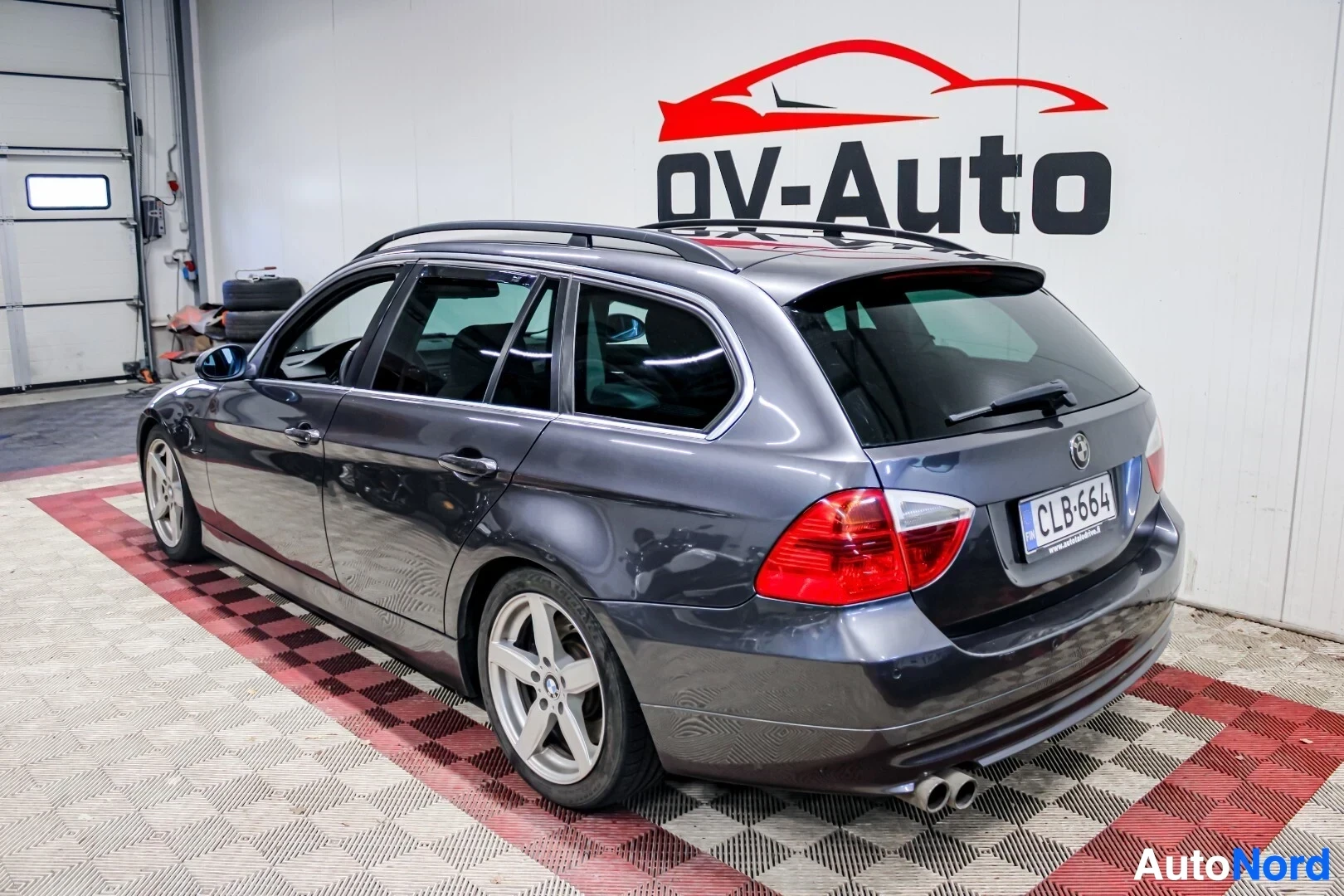 Takaviisto: Harmaa BMW 3-sarjan Touringin takaosa ja oikea kylki autohallissa. Autossa on kattokiskot ja tummennetut takalasit.