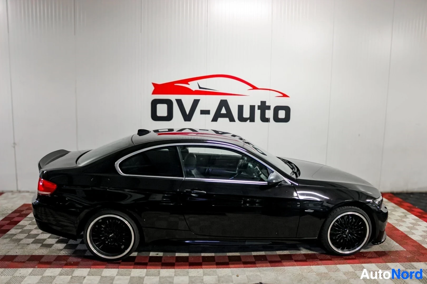 Sivulta: Musta coupe-auto pysäköitynä valkoisen seinän edessä, jossa on punainen ja valkoinen OV-Auto-logo. Auton sivuprofiili on selvästi näkyvissä.