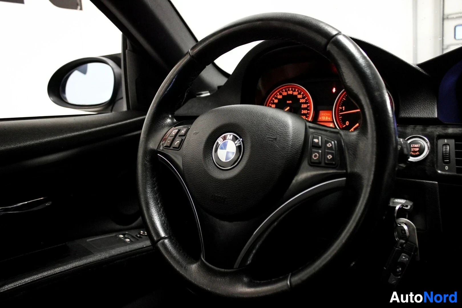 Yksityiskohta: Lähikuva auton ohjauspyörästä, jossa on BMW-logo ja monitoiminäppäimet. Taustalla näkyy mittaristo ja auton avaimet virtalukossa.