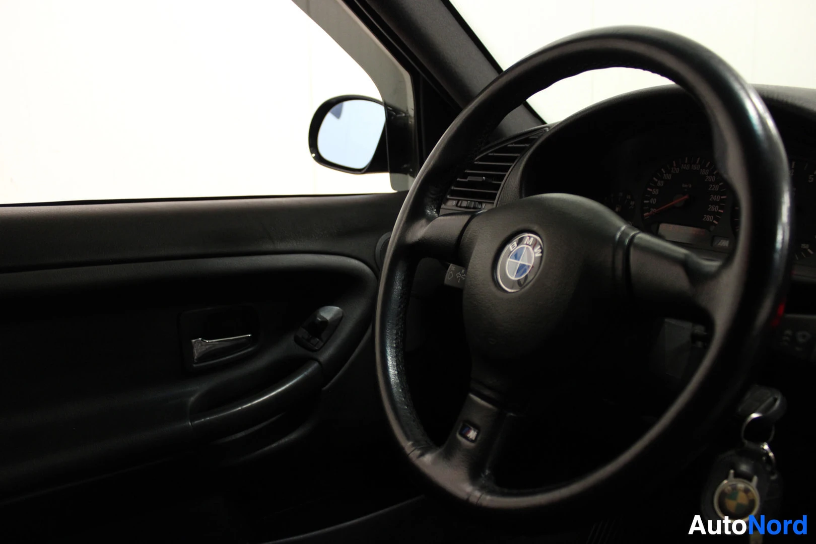 Ohjaamo: Kuva auton ohjaamosta, jossa näkyvät ohjauspyörä, mittaristo ja oven sisäpaneeli. Ohjauspyörän keskiössä on BMW-logo ja alareunassa M-merkki. Kuljettajan ovi ja