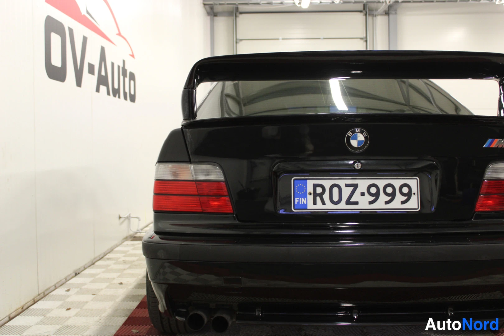 Takaa: Mustan auton takaosa lähikuvassa. Rekisterikilvessä lukee ja kilven yläpuolella näkyy BMW-logo sekä M-logo. Autossa on suuri takaspoileri ja kaksoispako