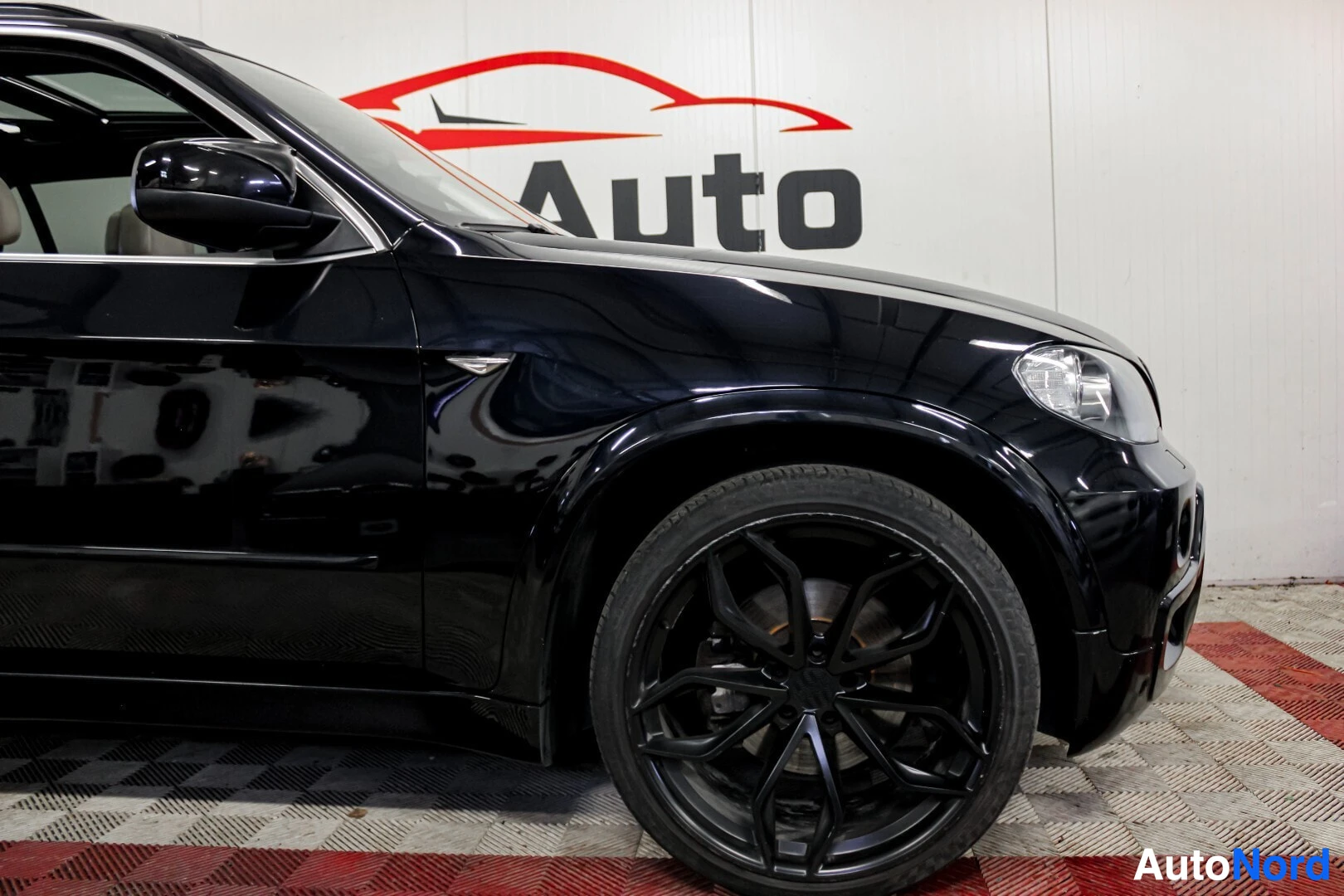 Etuviisto, nokka oikealle: Musta BMW X5:n etuosaa ja kylkeä esittävä kuva. Etulokasuoja ja ovi ovat näkyvissä. Autossa on kiiltävän mustat monipuolaiset kevytmetallivanteet ja mata