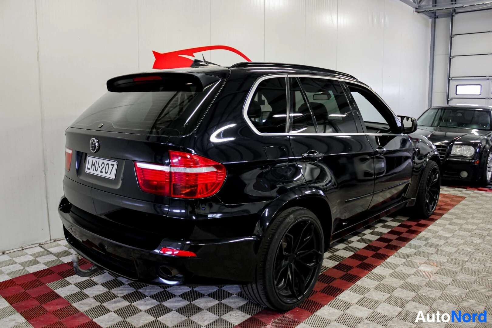 Takaviisto: Musta BMW X5 näkyy takaviistosta oikealta puolelta sisätiloissa. Autossa on mustat vanteet ja kattokaiteet.