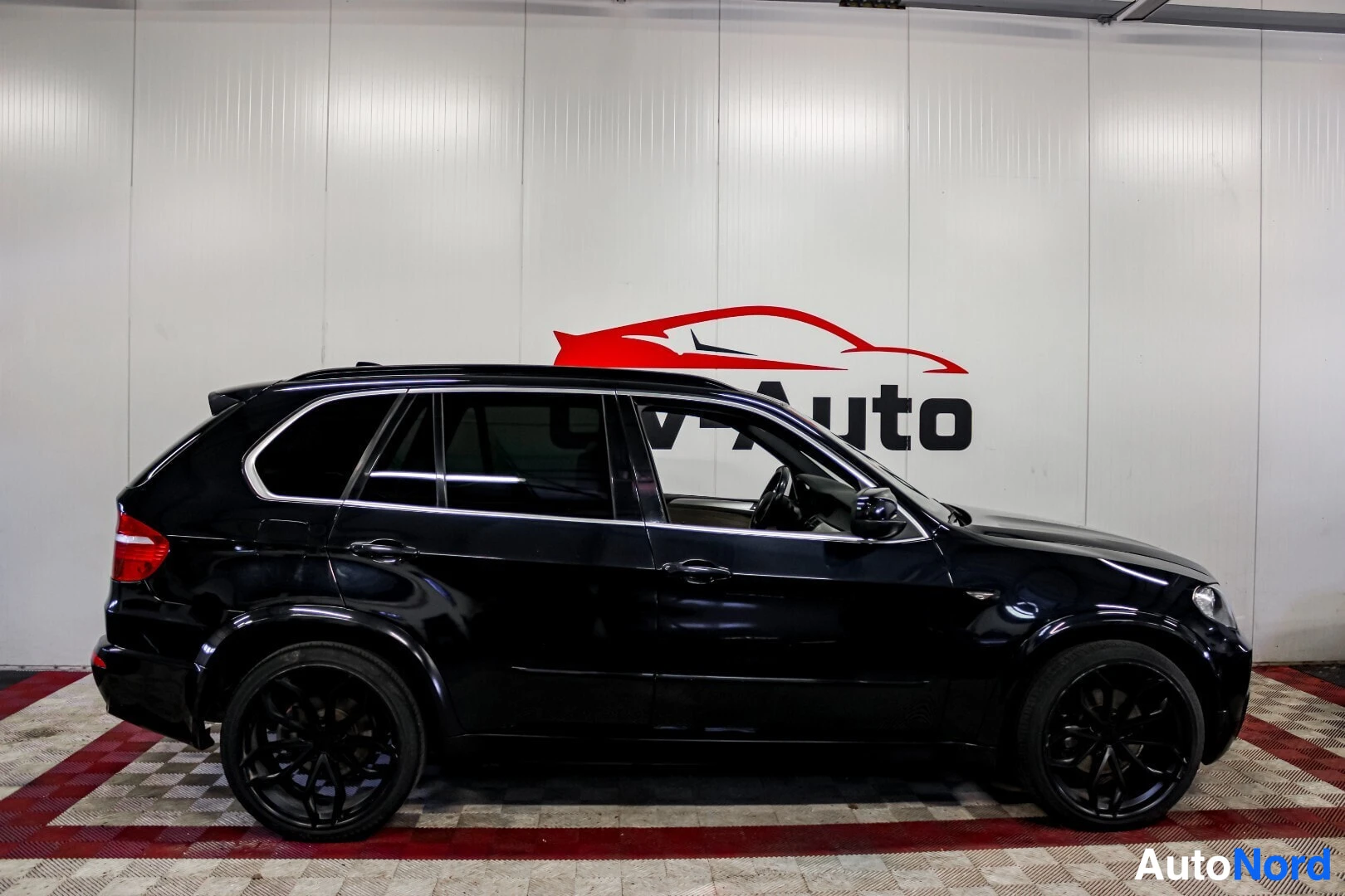 Sivulta: Musta BMW X5:n vasen sivu on kuvattu sisätiloissa. Autossa on mustat vanteet ja kattokaiteet.
