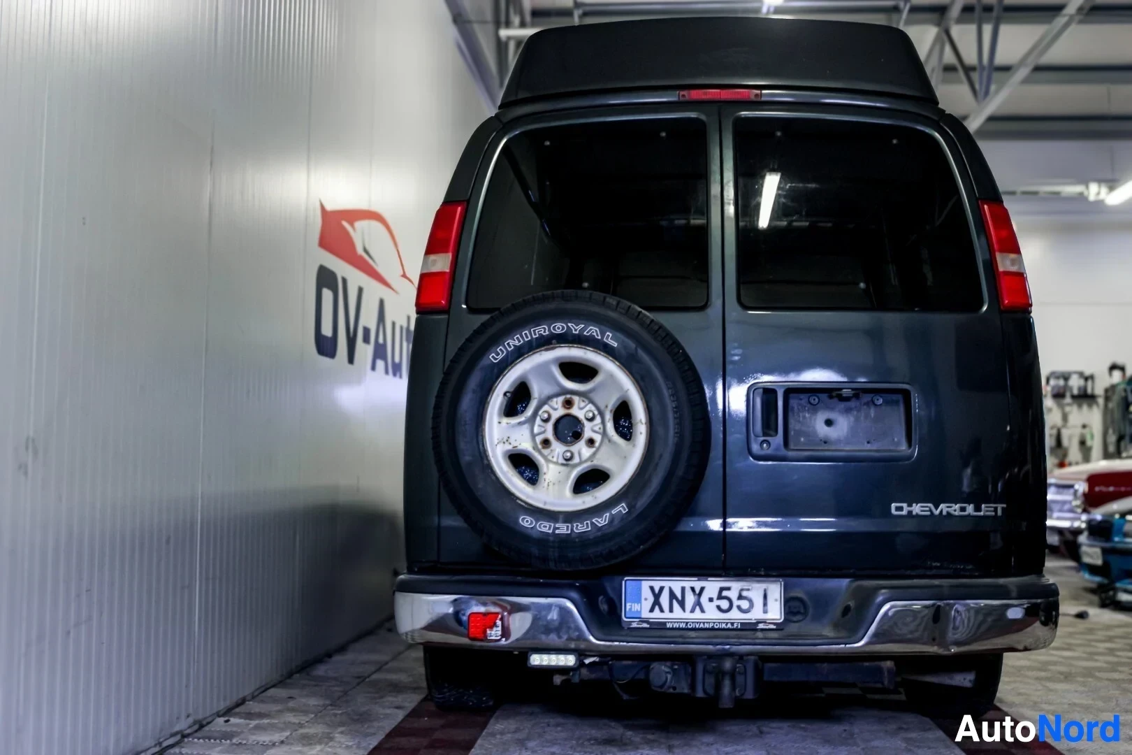 Takaa: Musta pakettiauto, jossa on korotettu katto, on kuvattu suoraan takaa. Auton takana on vararengas telineessä. Takapuskuri on kromattu ja sen yläpuolella on lisä