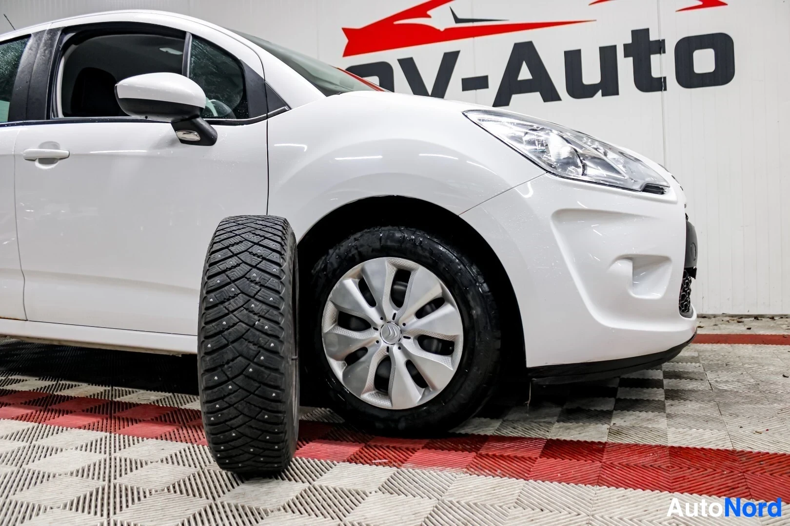 Etuviisto, nokka oikealle: Valkoinen auto pysäköitynä. Edessä on nastarengas. Auton oikea etupyörä on näkyvissä, vanteet ovat harmaat, renkaissa on nastat.