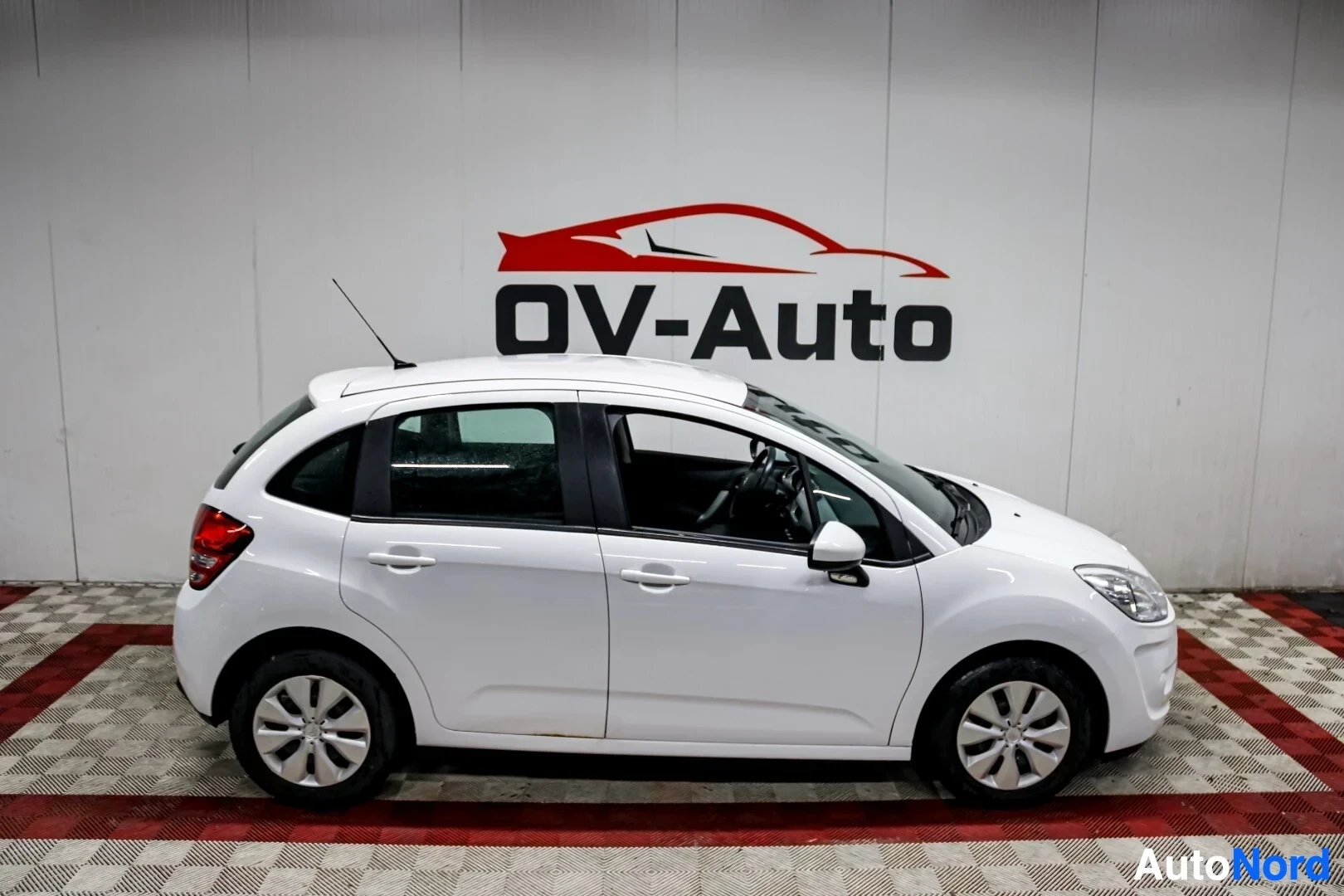 Sivulta: Valkoinen Citroen C3 vasemmalta sivulta kuvattuna, pysäköitynä halliin. Autossa on peltivanteet koristekapseleilla.