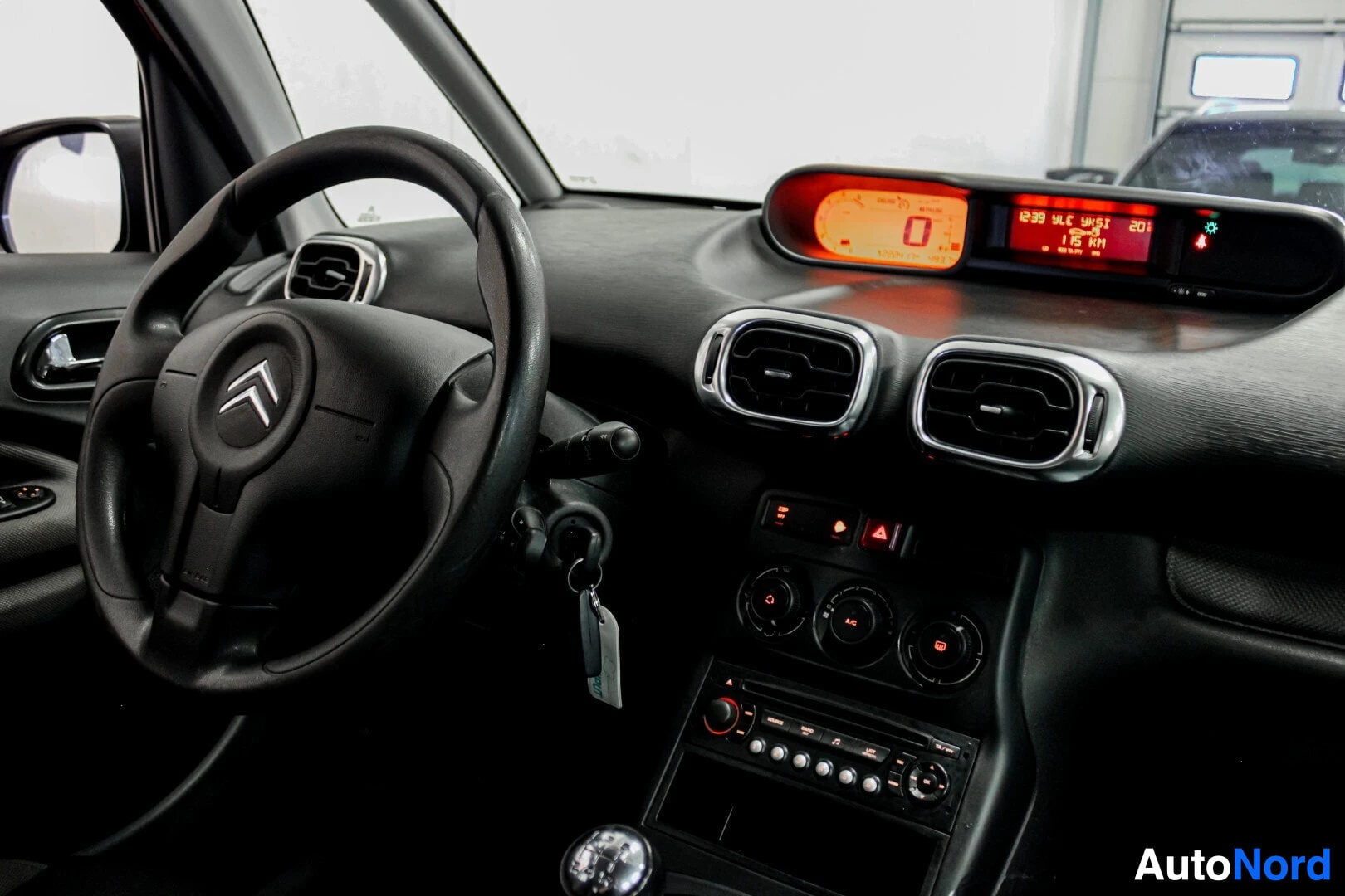 Ohjaamo: Kojelauta ja ohjauspyörä, jossa Citroënin logo, edessä kuljettajan paikka. Kojelaudalla näkyy oranssipohjainen digitaalinen näyttö.