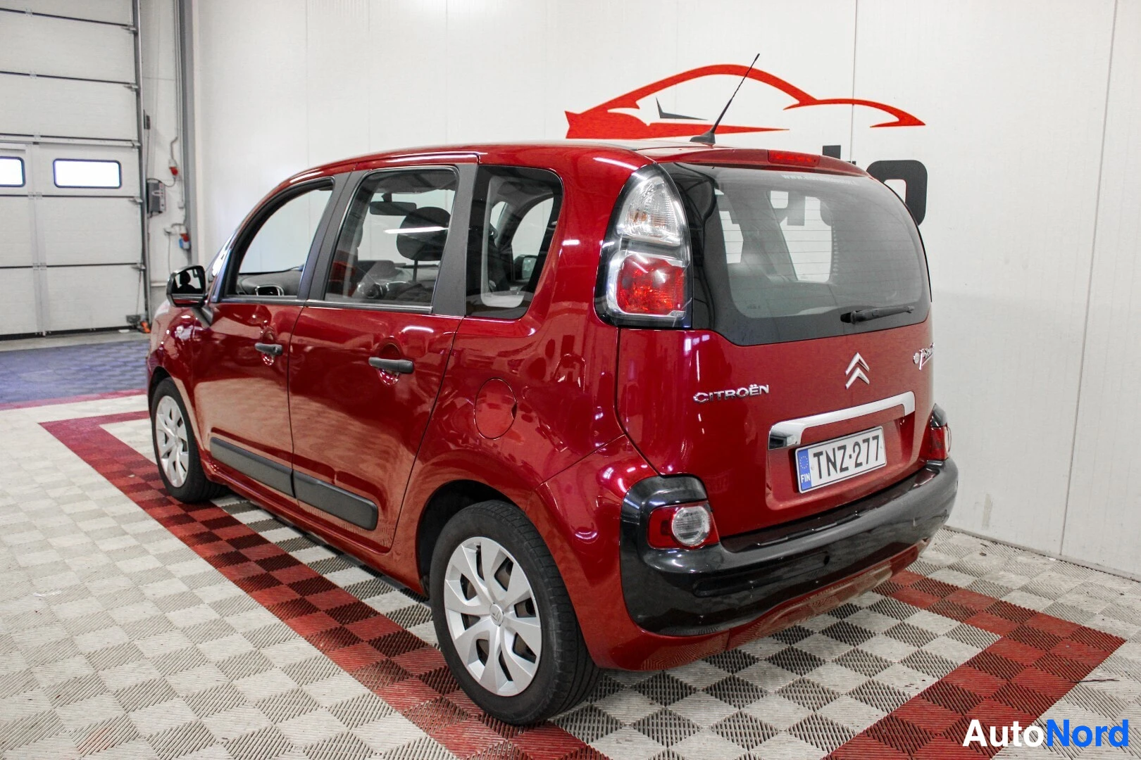 Takaviisto: Punainen Citroën C3 Picasso seisoo salissa, sen takaosa ja vasen kylki näkyvät takavasemmalta kuvattuna. Rekisterikilpi on