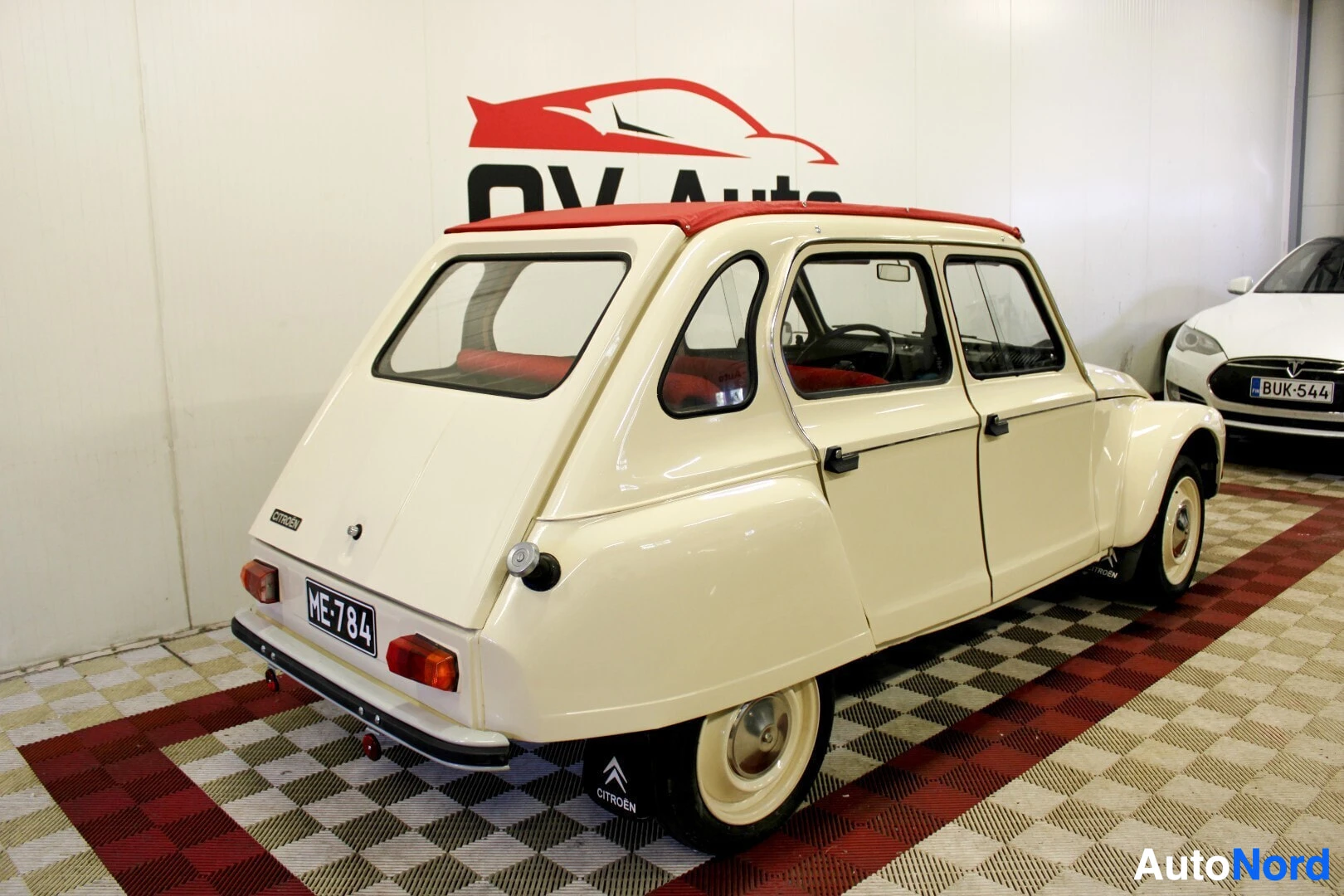 Takaviisto: Beige auto punaisella kangaskatolla kuvattuna takavasemmalta viistoon sisätilassa. Takarekisterikilpi ME-784 näkyy. Oikealla takana näkyy osittain toinen valkoi