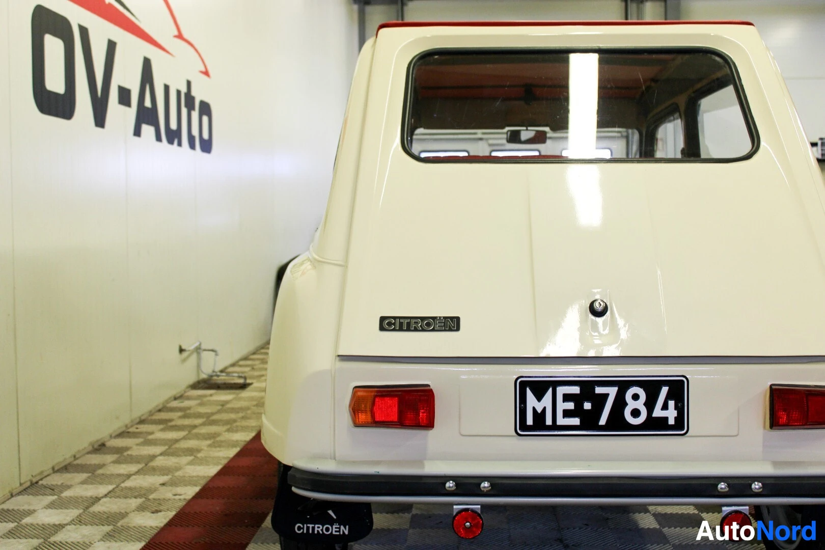 Takaa: Beige auton takaosa suoraan takaa kuvattuna. Rekisterikilpi ME-784 näkyy selkeästi. Vasemmalla näkyy seinä, jossa lukee osittain "OV-Auto".