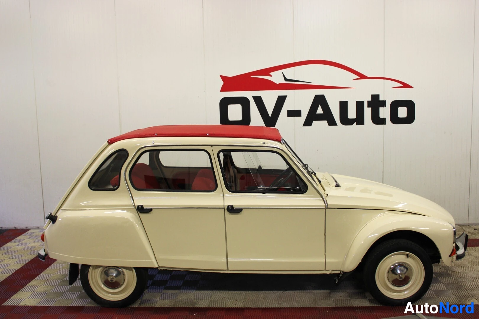 Sivulta: Auton vasen kylki kuvattuna auton sivusta. Autossa on vaaleanbeige kori ja punainen kangaskatto. Edessä on valkoinen seinä, jossa on punamusta "OV-Auto"-logo.