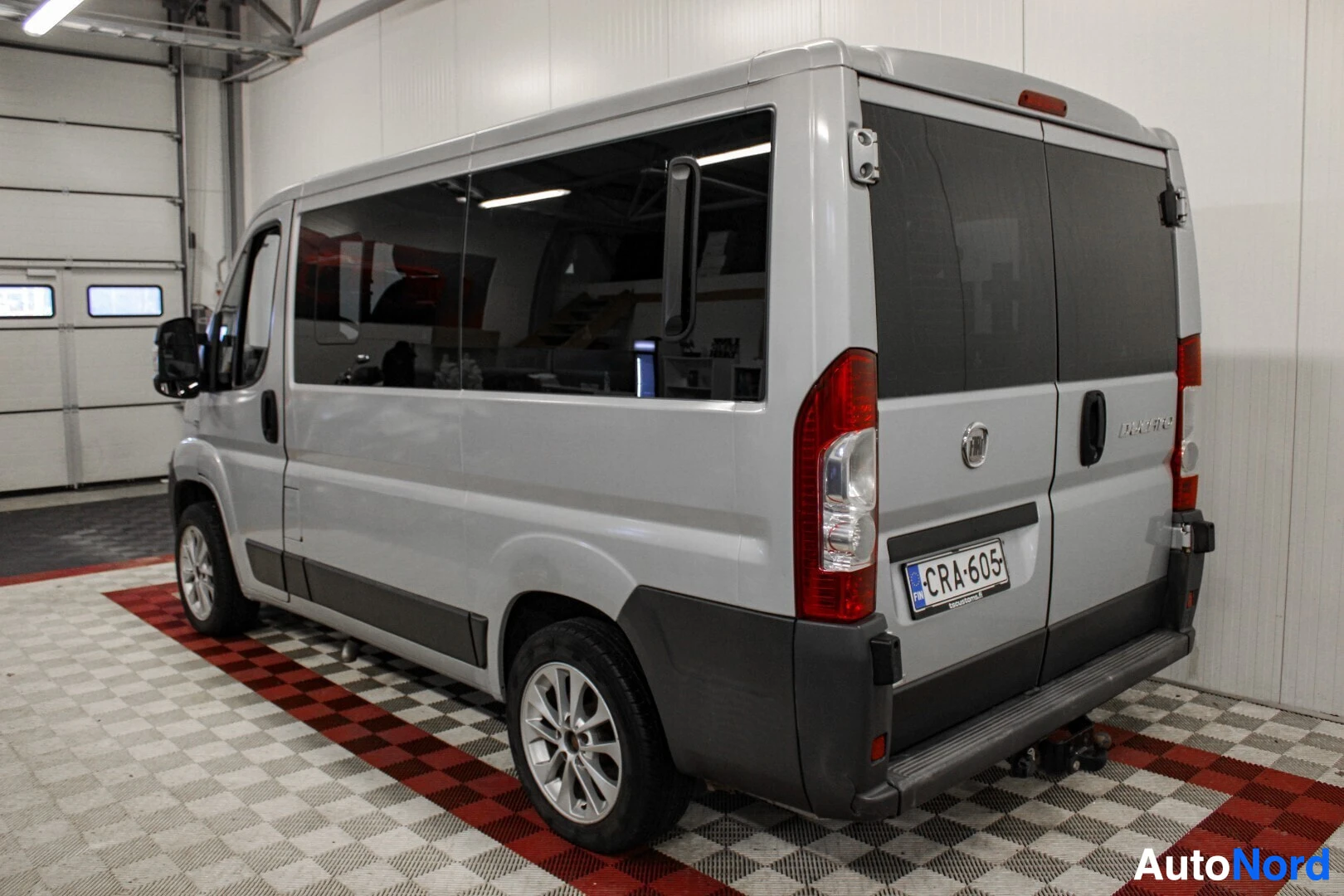 Takaviisto: Hopeanvärinen pakettiauto, jossa on vetokoukun pistoke takana ja alumiinivanteet, pysäköity sisätiloihin shakkiruutuisella lattialla.