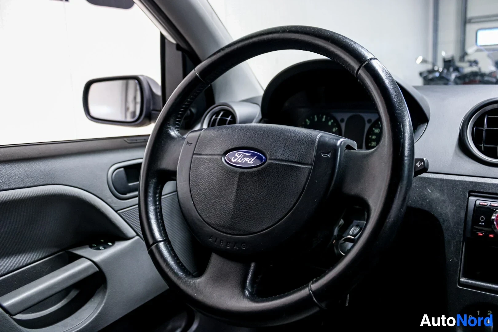 Ohjaamo: Kuvakulma auton ohjaamosta, jossa näkyy nahkainen ohjauspyörä Ford-logolla ja "AIRBAG"-tekstillä. Taustalla näkyy osittain mittaristo.
