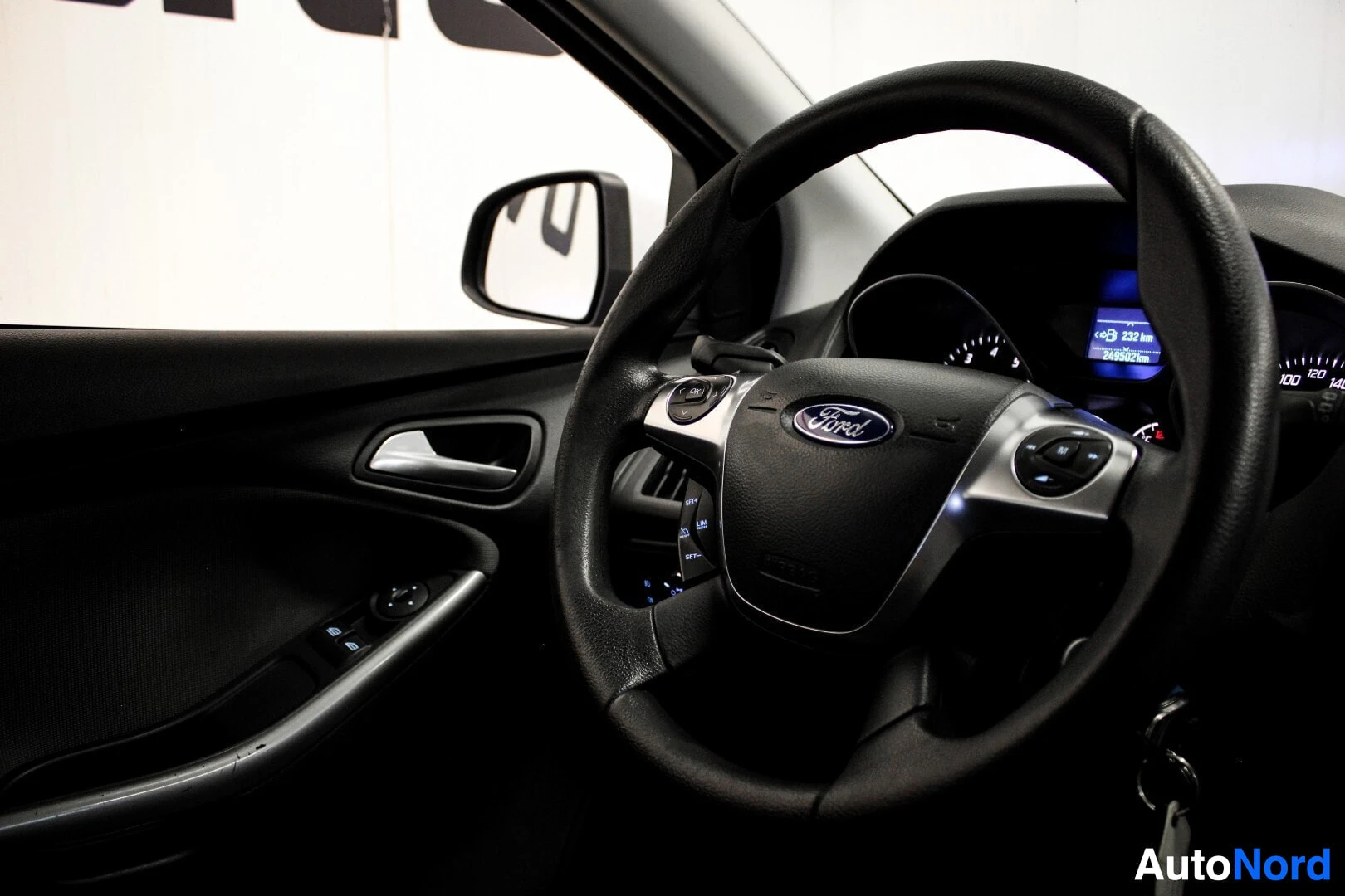 Ohjaamo: Kuva auton sisätiloista, jossa näkyvät ohjauspyörä, kuljettajan puoleinen ovenkahva ja ikkunoiden katkaisimet. Ohjauspyörässä on monitoiminäppäimet ja Ford-logo