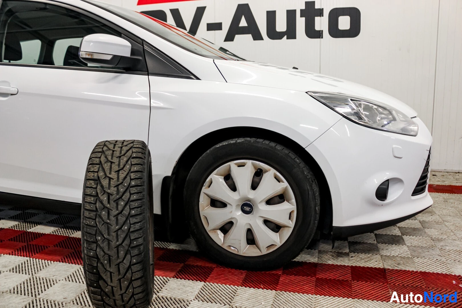 Etuviisto, nokka vasemmalle: Valkoinen auto edestä ja oikealta sivulta. Edessä oikea ajovalo ja etupuskuri sumuvalon aukolla. Oikea eturengas on kuvassa ja sen vieressä pystyssä na