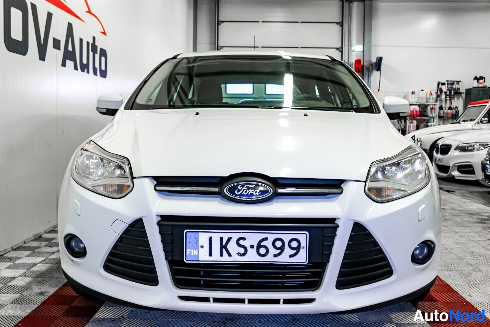 Edestä: Valkoinen farmariauto suoraan edestä kuvattuna parkkihallissa. Autossa on Ford-logo jäähdyttäjän säleikössä ja rekisterinumero