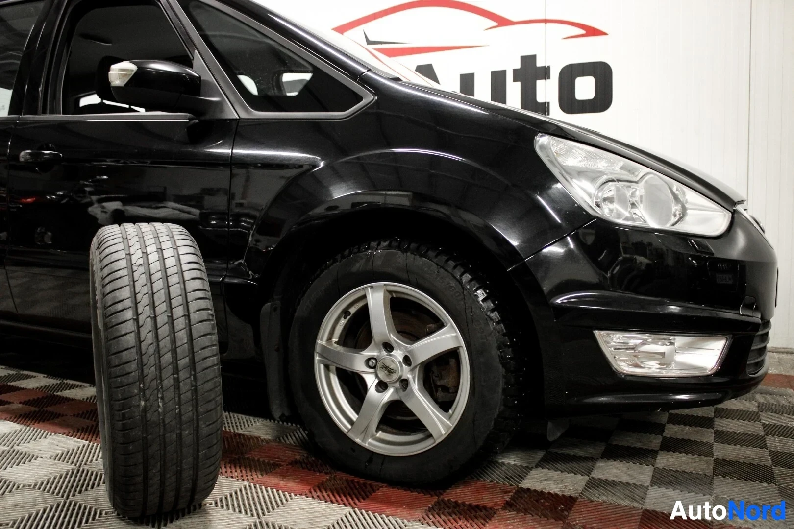 Etuviisto, nokka vasemmalle: Musta auto edestä oikealta, auton vieressä vanteeton kesärengas ja autossa talvirengas alumiinivanteella.