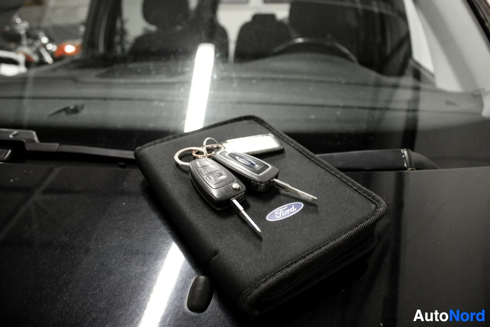 Yksityiskohta: Kaksi Ford-logoista auton avainta ja ohjekirjan tapainen kansio mustan auton konepellillä.