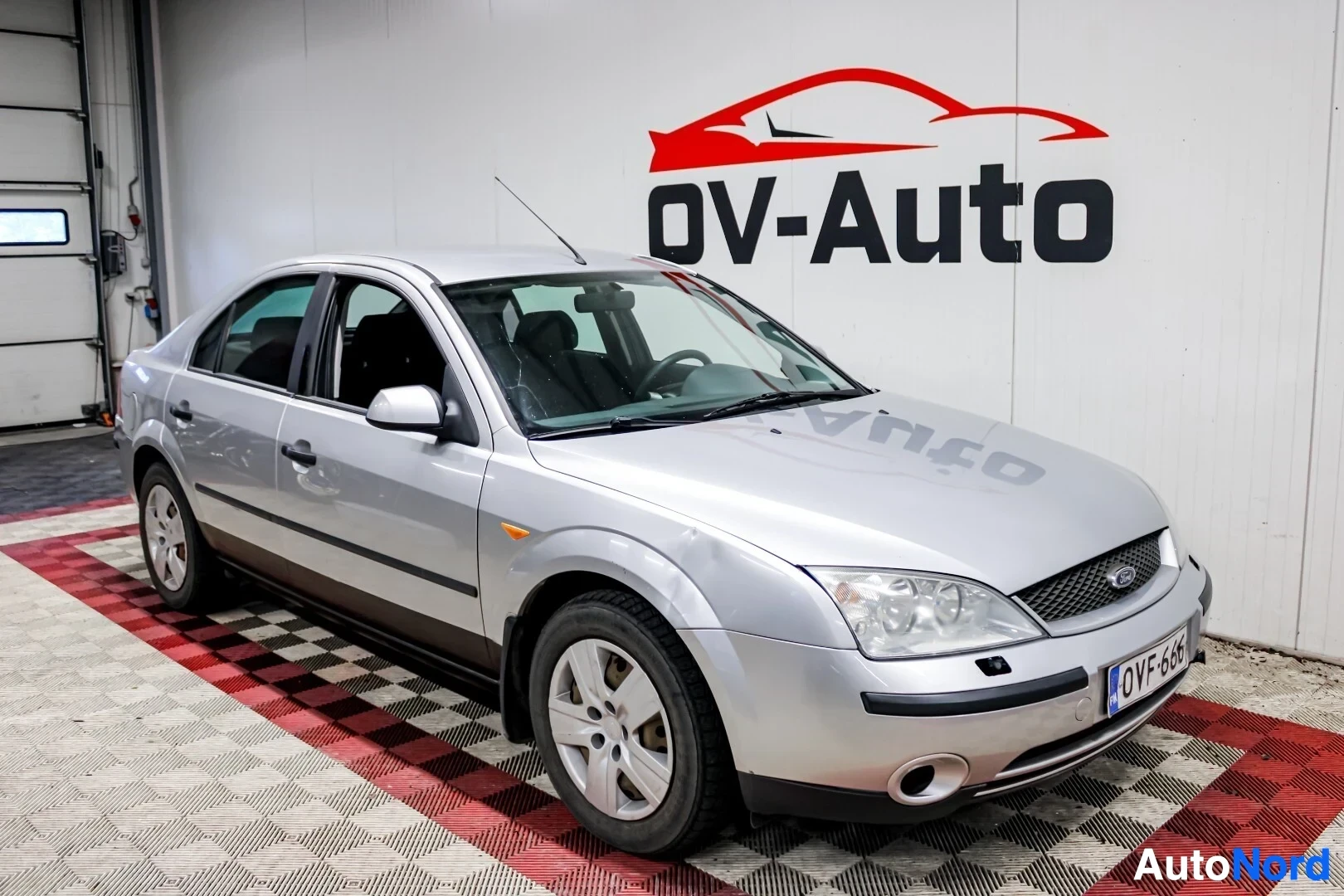 Etuviisto, nokka oikealle: Hopeanvärinen Ford Mondeo on pysäköity halliin, etu- ja sivukulma kuvattuna. Auton takana on OV-Auto-logo. Autossa on musta eturekisterikilpi