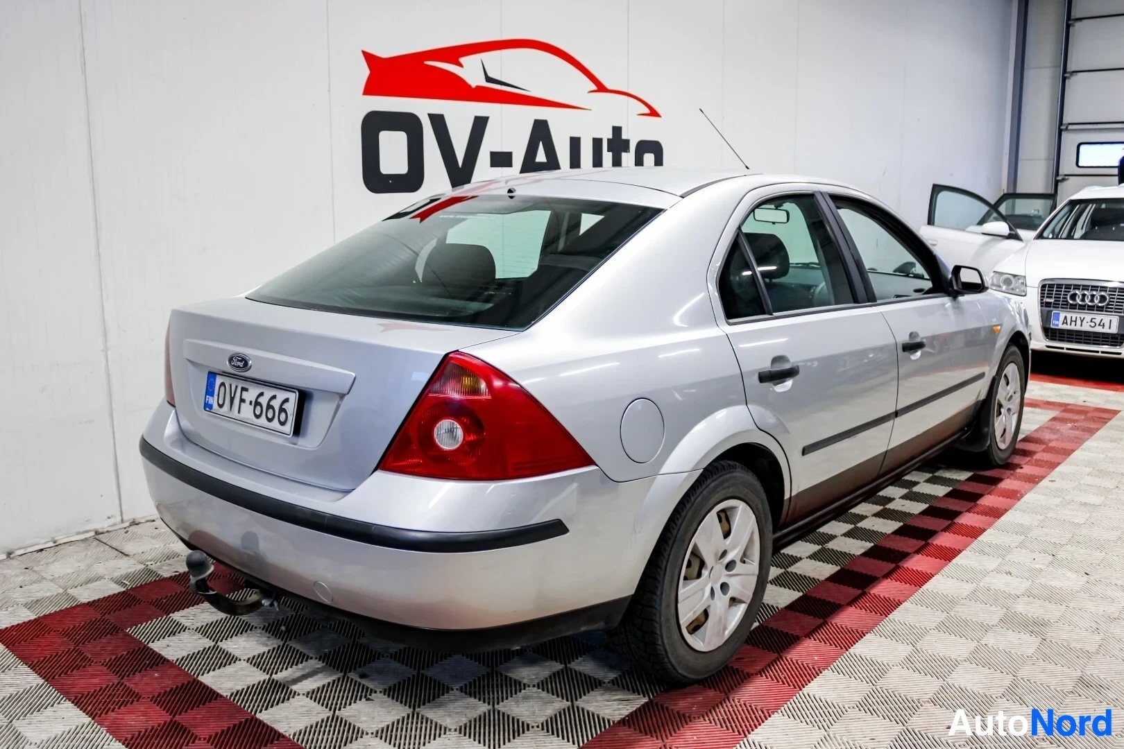 Takaviisto: Hopeanvärinen Ford Mondeo henkilöauto takavasemmalta kuvattuna OV-Auto-logon edessä. Ajoneuvon takavalot, rekisterikilpi, vetokoukku ja vasen kylki ovat
