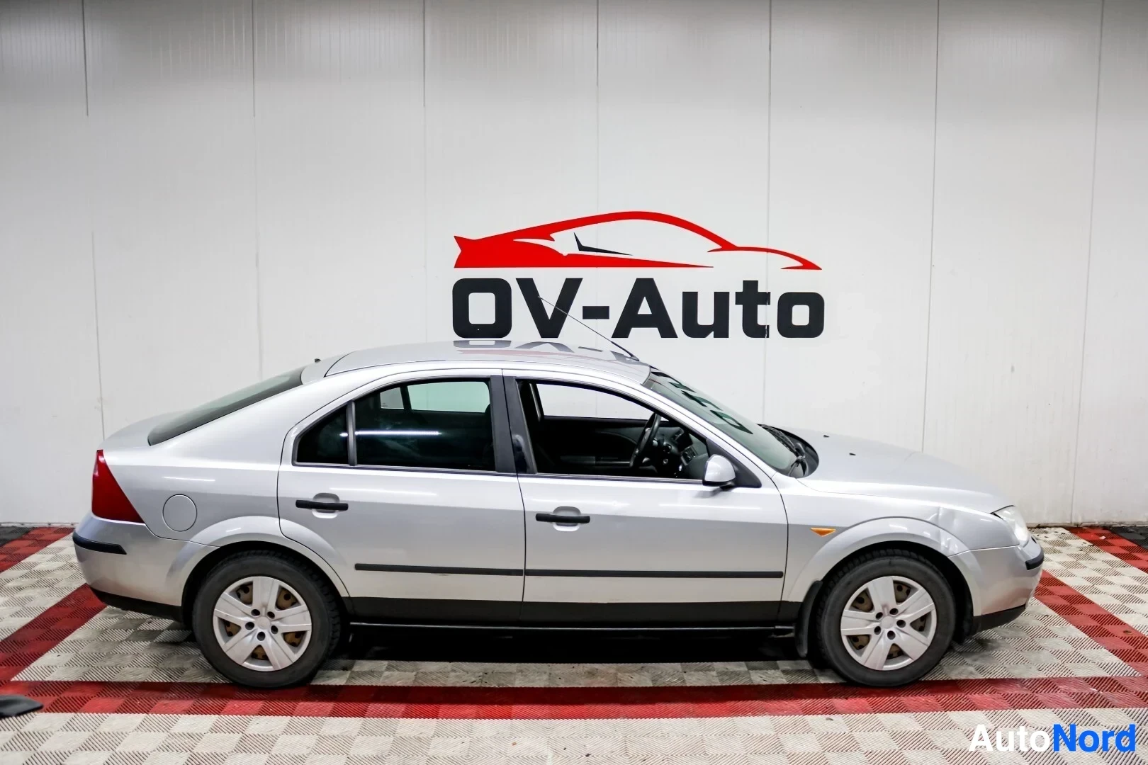 Sivulta: Hopeanvärinen Ford Mondeo henkilöauto kuvattuna suoraan sivulta vasemmalta. Auto on pysäköity OV-Auto-logon eteen.