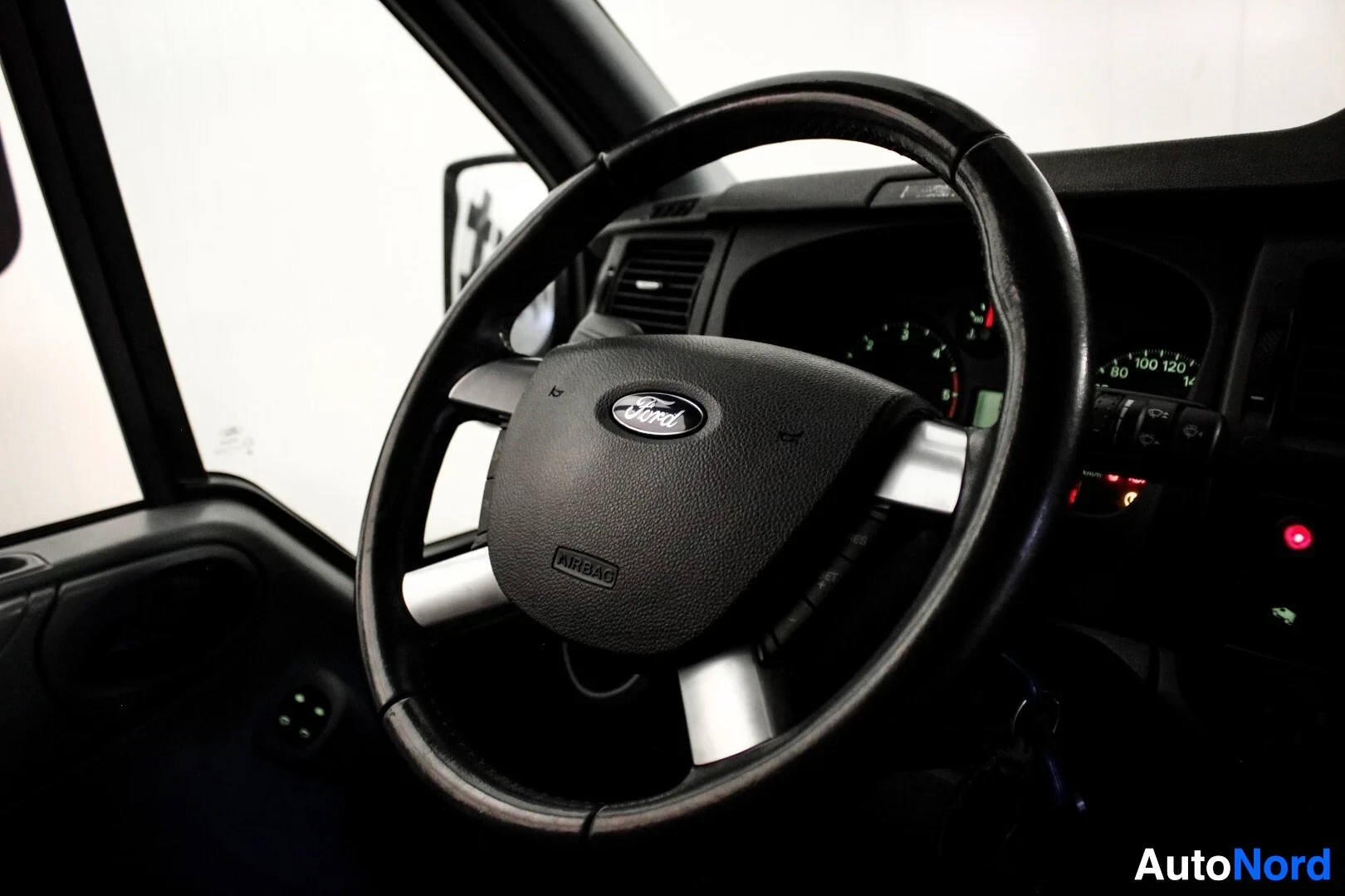Ohjaamo: Musta kolmipuolainen ohjauspyörä auton sisätilassa. Keskellä on Ford-logo ja sana "AIRBAG". Ohjauspyörän takana näkyy mittaristo ja ovenkahvan vieressä kaksi sä