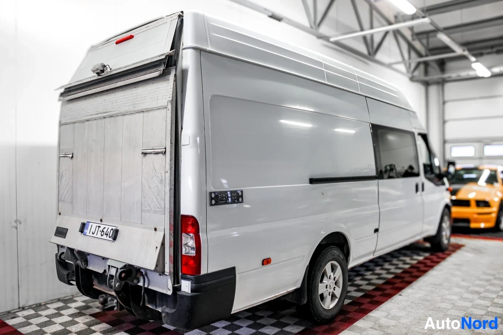Takaviisto: Valkoisen pakettiauton takaosa ja oikea kylki kuvattuna vinosti takaapäin. Tavaratilan ovi on auki ja sen takana näkyy mustakeltainen auto. Rekisterikilpi IJ T-