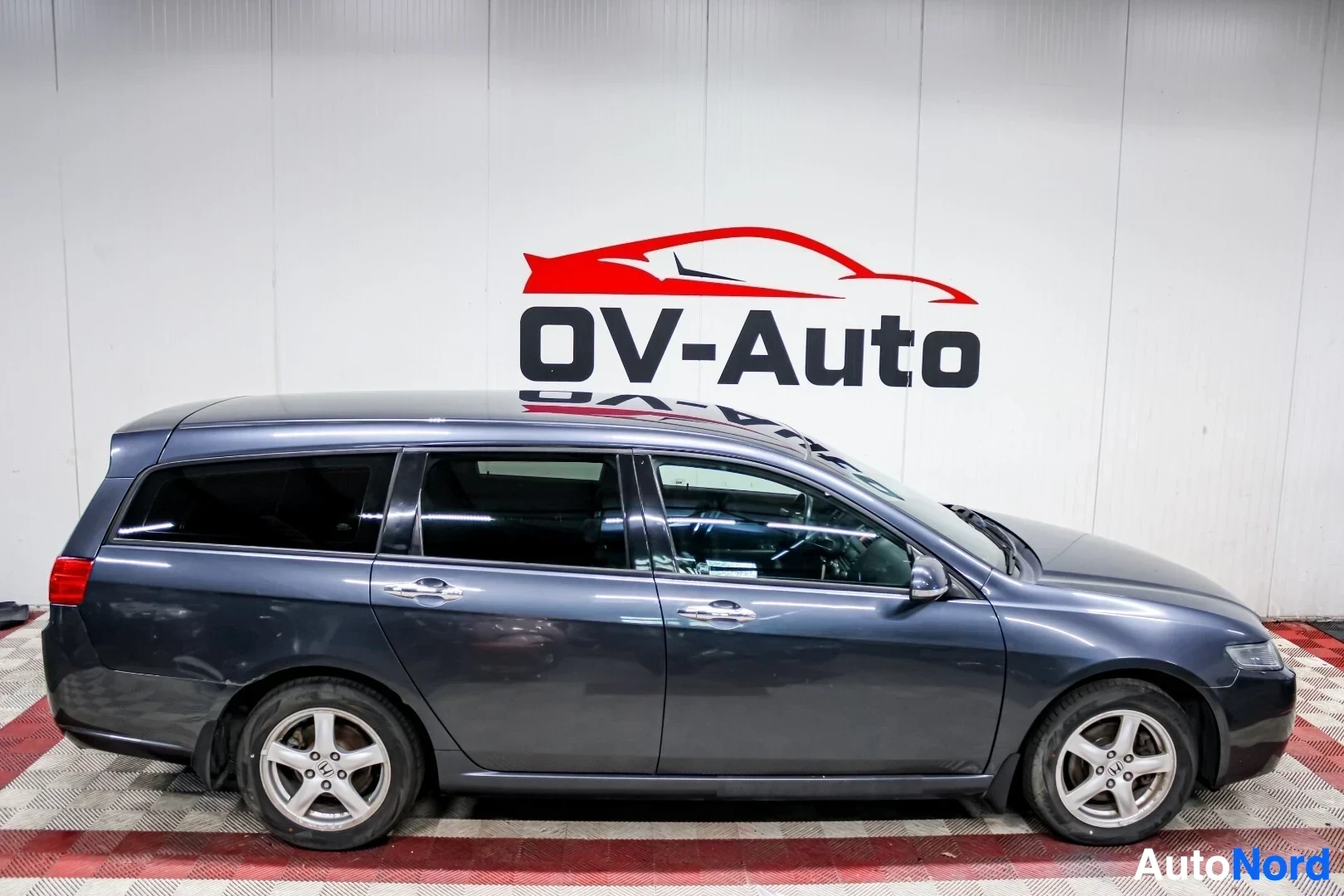 Sivulta: Tummanharmaa Honda Accord Tourer nähtynä suoraan sivulta. Auto on pysäköity sisätiloihin, ja taustalla on valkoinen seinä, jossa on autoliikkeen logo. Molemmat