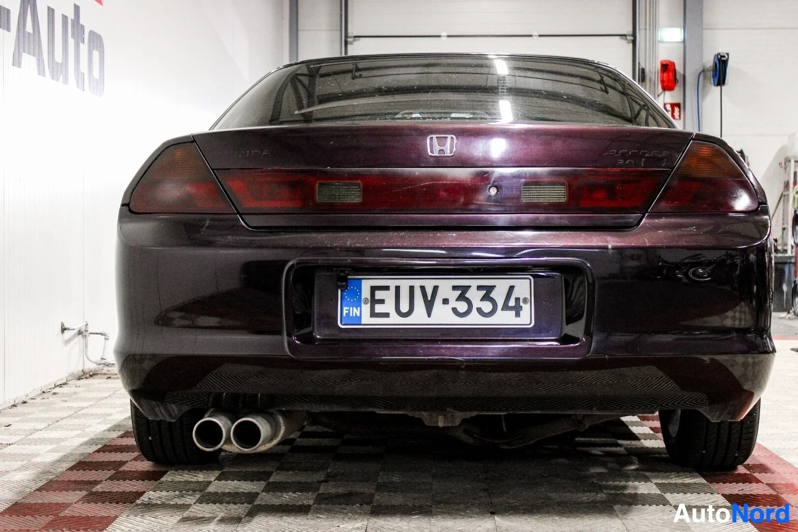 Takaa: Tummanvioletti coupé-auto kuvattuna suoraan takaa OV-Auton logolla varustetun seinän edessä. Rekisterikilpi on näkyvissä ja ajoneuvossa on kaksi pakoput