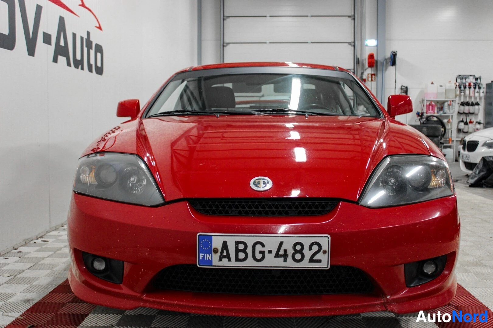 Edestä: Punaisen coupé-auton etuosa ruudullisella lattialla sisätiloissa. Rekisterikilpi, jossa lukee "FIN ", on näkyvissä edessä