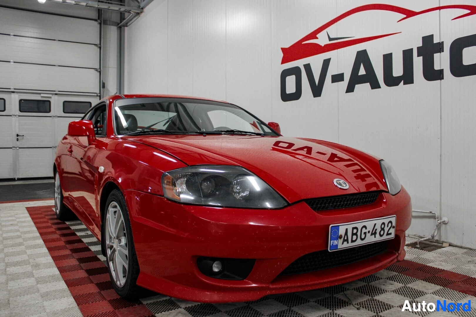 Etuviisto, nokka oikealle: Punainen coupé-auto pysäköitynä ruudulliselle lattialle sisätiloissa, etuvasen kulma näkyvissä. Auton edessä näkyy rekisterikilpi tekstillä "FIN "