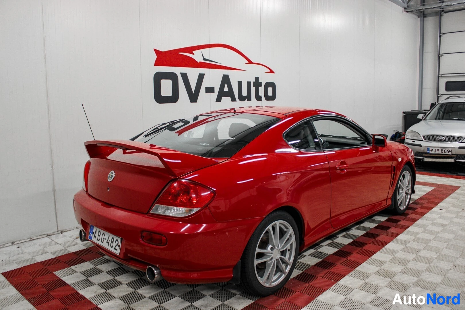 Takaviisto: Punainen kaksiovinen coupé-auto takaoikealta kuvattuna. Auton takana on valkoinen seinä, jossa lukee mustalla tekstillä "OV-Auto" ja punaisella auton siluetilla