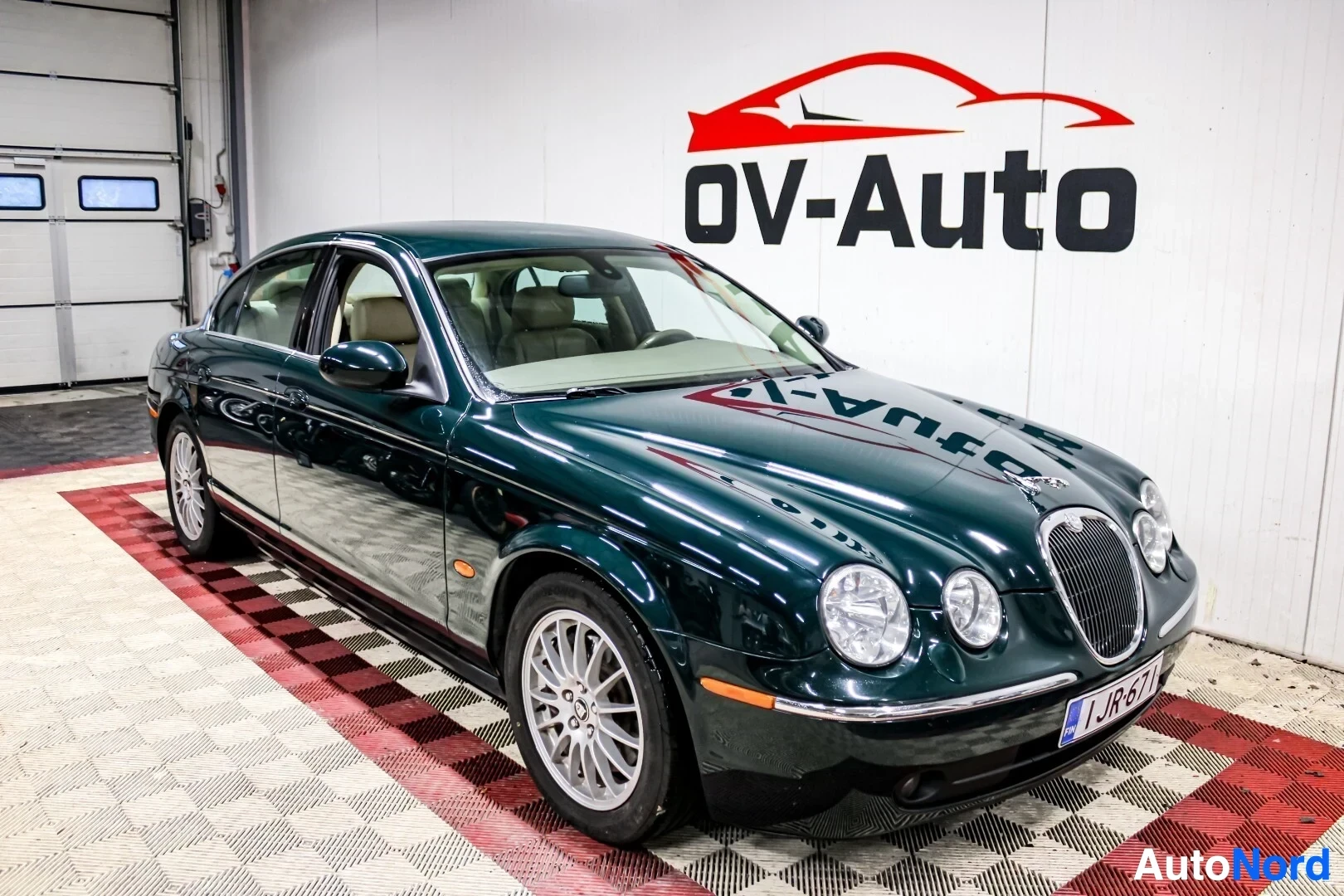 Etuviisto, nokka vasemmalle: Tummanvihreä Jaguar S-Type sedan seisoo valkoisella ja punaisella ruudullisella lattialla autokaupan hallissa. Auton vasen etu- ja kylkiosat ovat kuvas