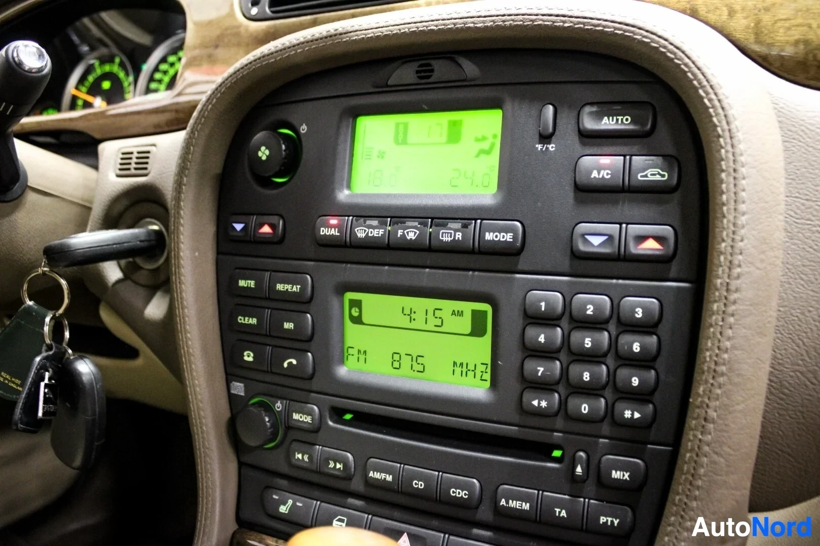 Ohjaamo: Lähikuva auton keskikonsolista, jossa on radio ja ilmastoinnin säätimet, molemmissa vihreä digitaalinäyttö. Radion näytöllä lukee "FM 87.5 MHZ" ja "4:15". Ilmas
