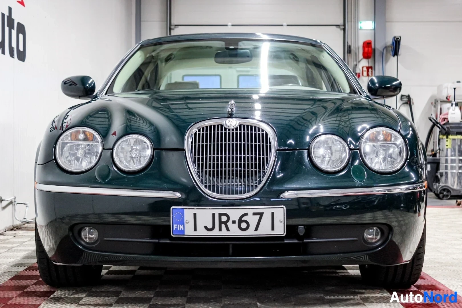Edestä: Tummanvihreän Jaguar S-Type -sedanin keula. Auton keskellä on pystysuuntainen säleikkö ja sen yläpuolella Jaguar-logo. Molemmin puolin on kaksi pyöreää ajovaloa