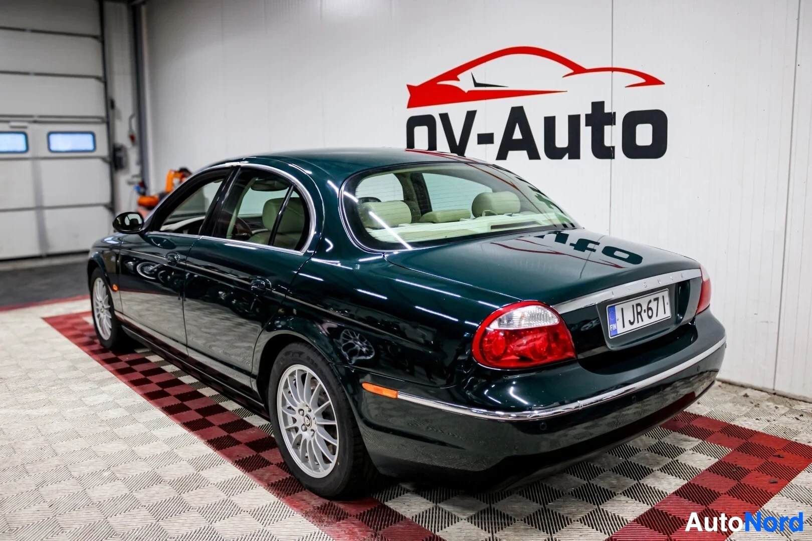 Takaviisto: Tummanvihreä Jaguar S-Type sedan seisoo valkoisella ja punaisella ruudullisella lattialla autokaupan hallissa. Auton oikea taka- ja kylkiosat ovat kuvassa. Valk