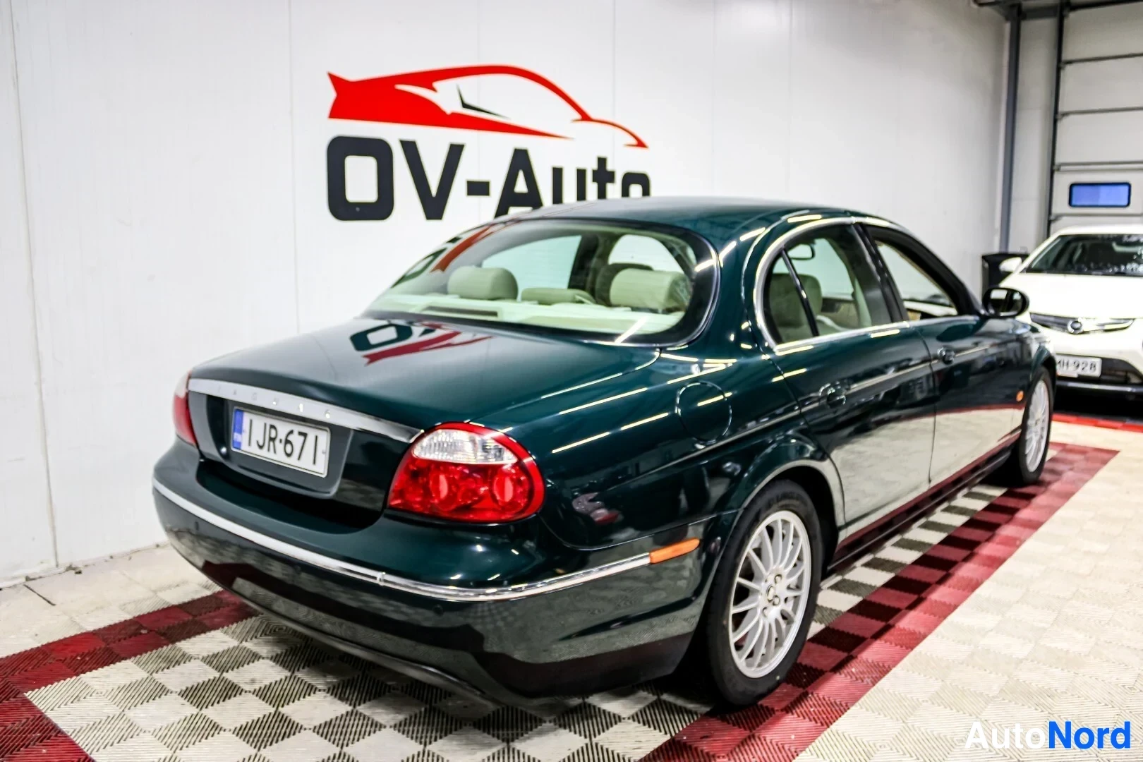 Takaviisto: Tummanvihreä Jaguar S-Type rekisterikilvellä pysäköitynä valkoisen ja punaisen OV-Auto-logon edessä. Takavalot ja ajovalot näkyvissä, sekä auton takaosa