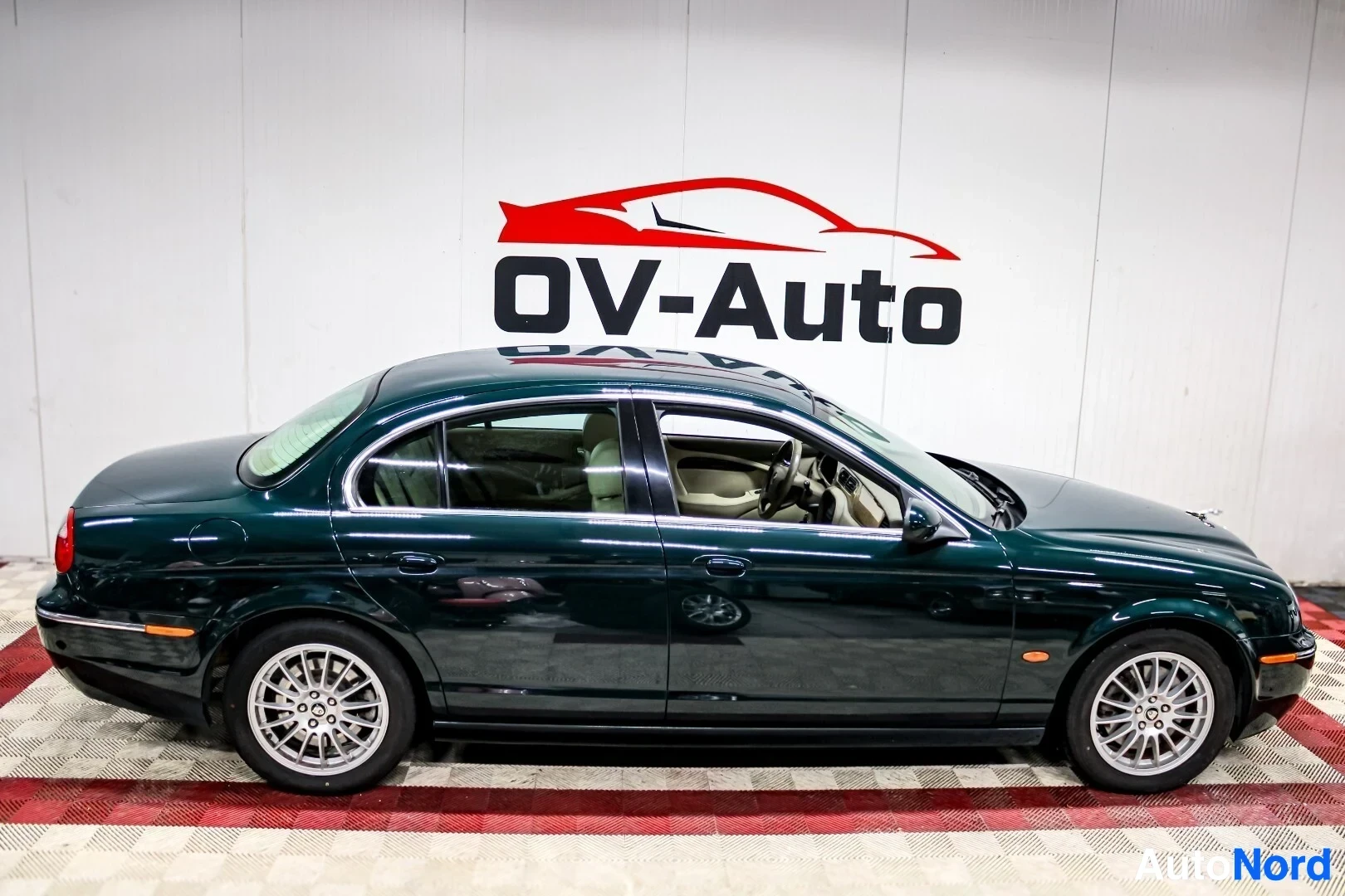 Sivulta: Tummanvihreä Jaguar S-Type pysäköitynä valkoisen ja punaisen OV-Auto-logon edessä, auton vasen kylki näkyvissä. Ikkunat ovat tummennetut ja valkoiset kevytmetal