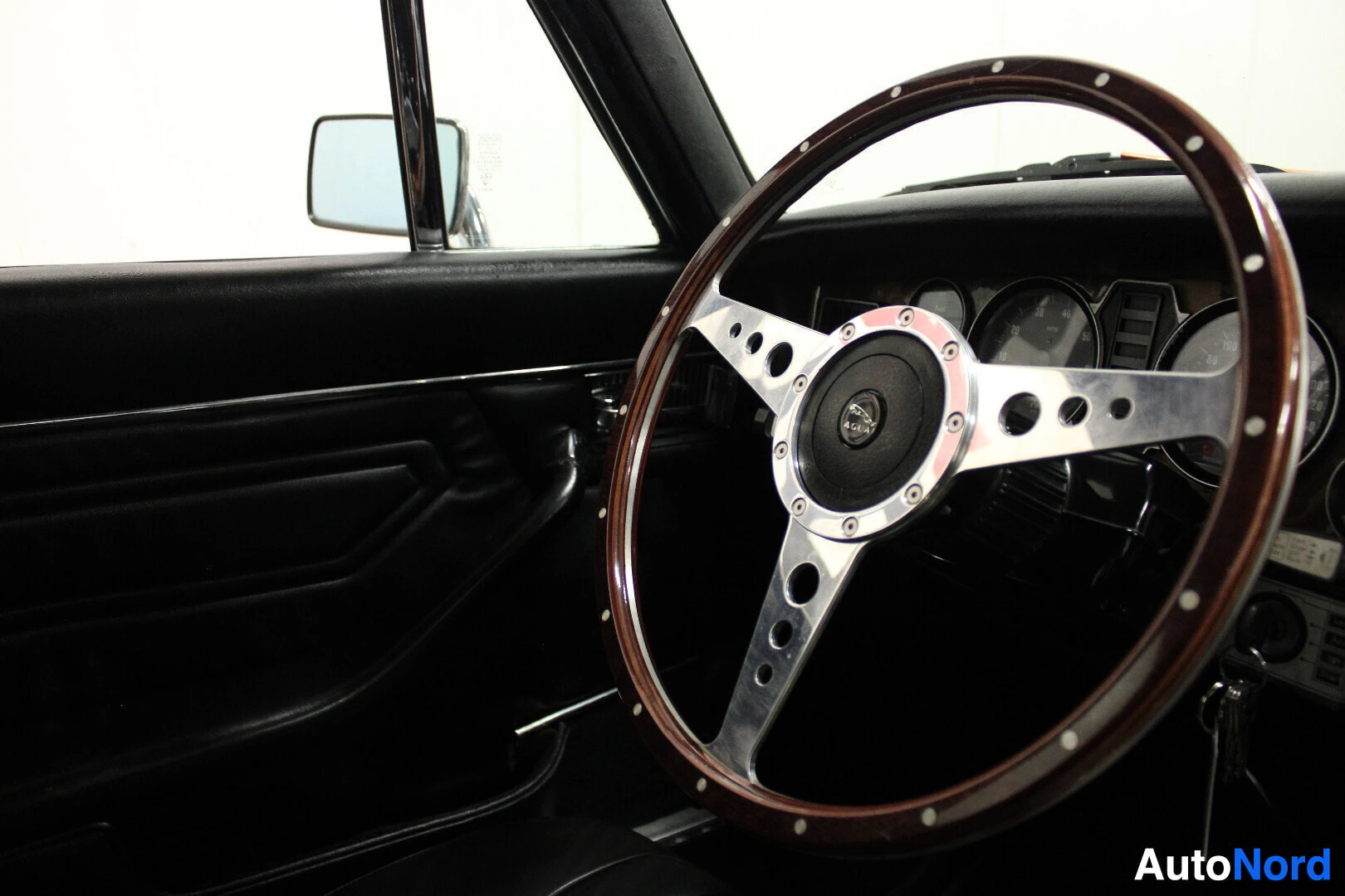 Ohjaamo: Lähikuva auton sisätiloista, etualalla kolmipuolainen puinen ohjauspyörä, jossa on reikiä metallisissa puolissa. Vasemmalla näkyy musta oven sisäpaneeli ja sivu