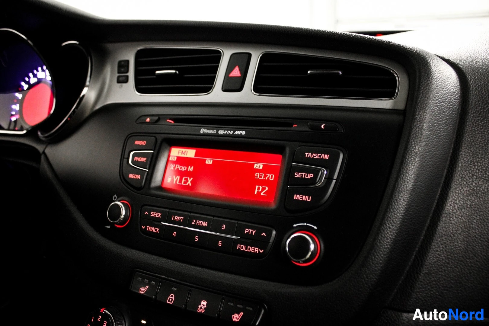 Yksityiskohta: Lähikuva auton keskikonsolista, jossa näkyy audiojärjestelmä punaisella näytöllä. Näytöllä lukee "YLEX" ja radiotaajuus "93.70". Yläpuolella on ilmanvaihtoaukot