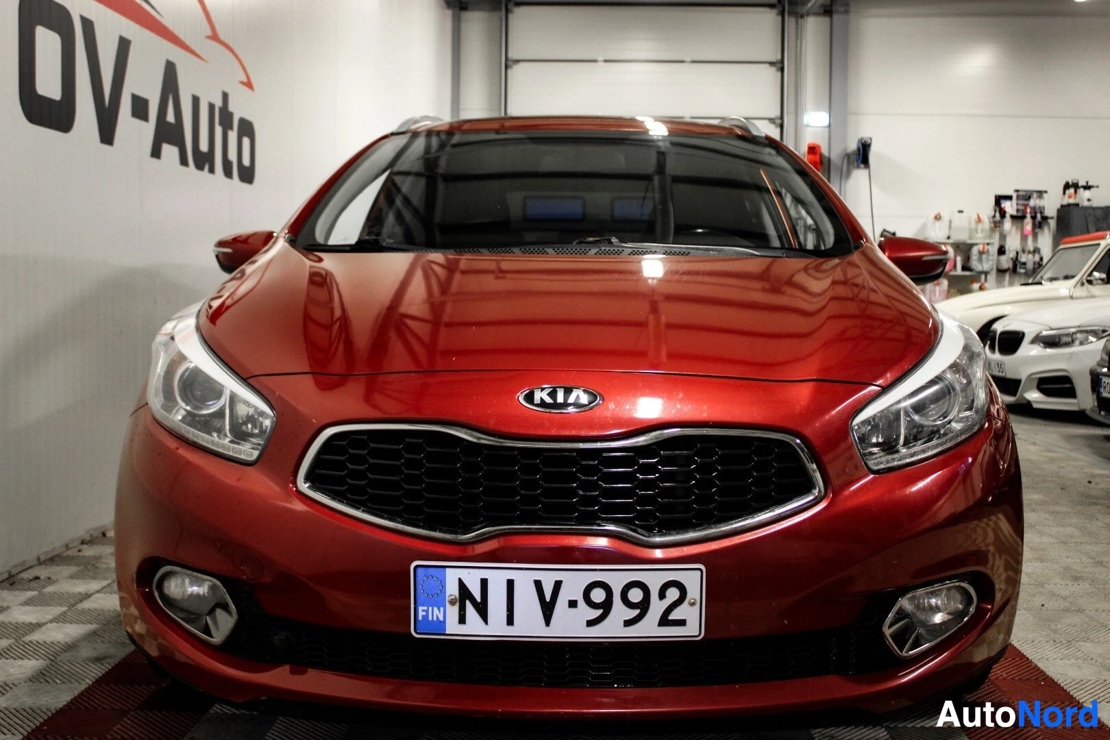 Edestä: Punaisen Kia Ceed farmarimallin keula edestä kuvattuna. Rekisterikilpi on kiinnitetty etusäleikön alapuolelle. Autossa on päiväajovalot ja sumuvalot.