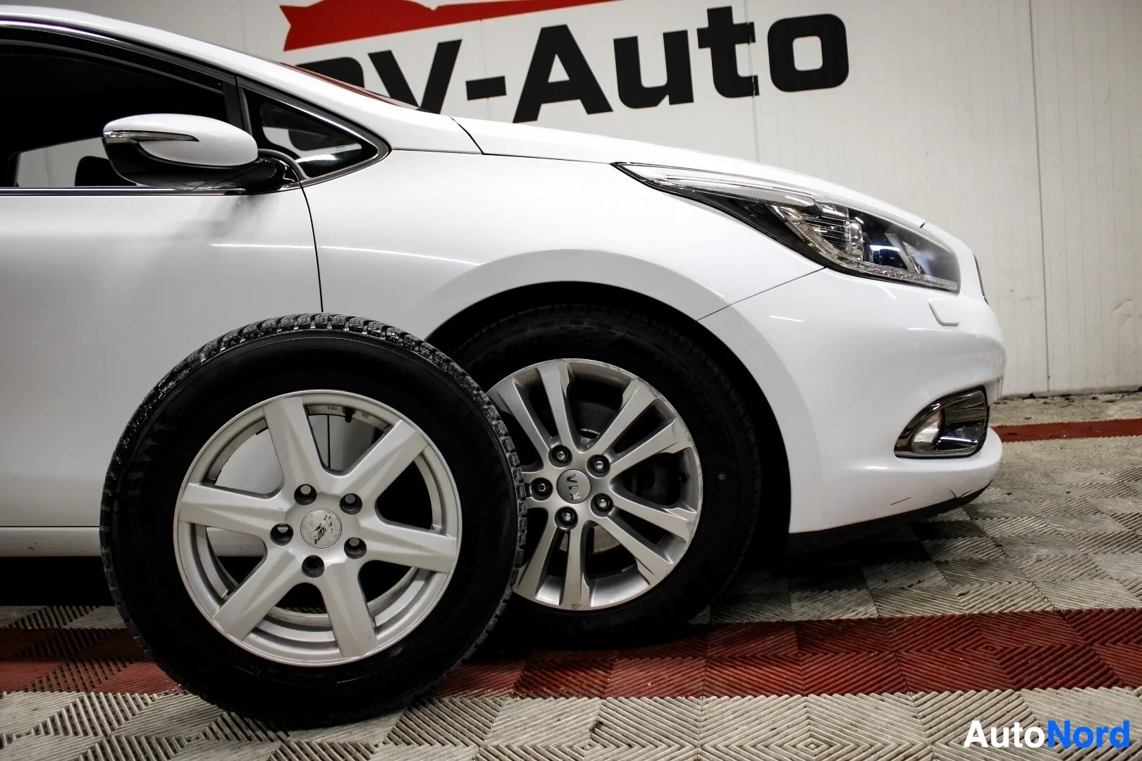 Etuviisto, nokka oikealle: Edestä viistoon kuvattu valkoinen henkilöauto. Auton edessä on irtonainen talvirengas alumiinivanteella.