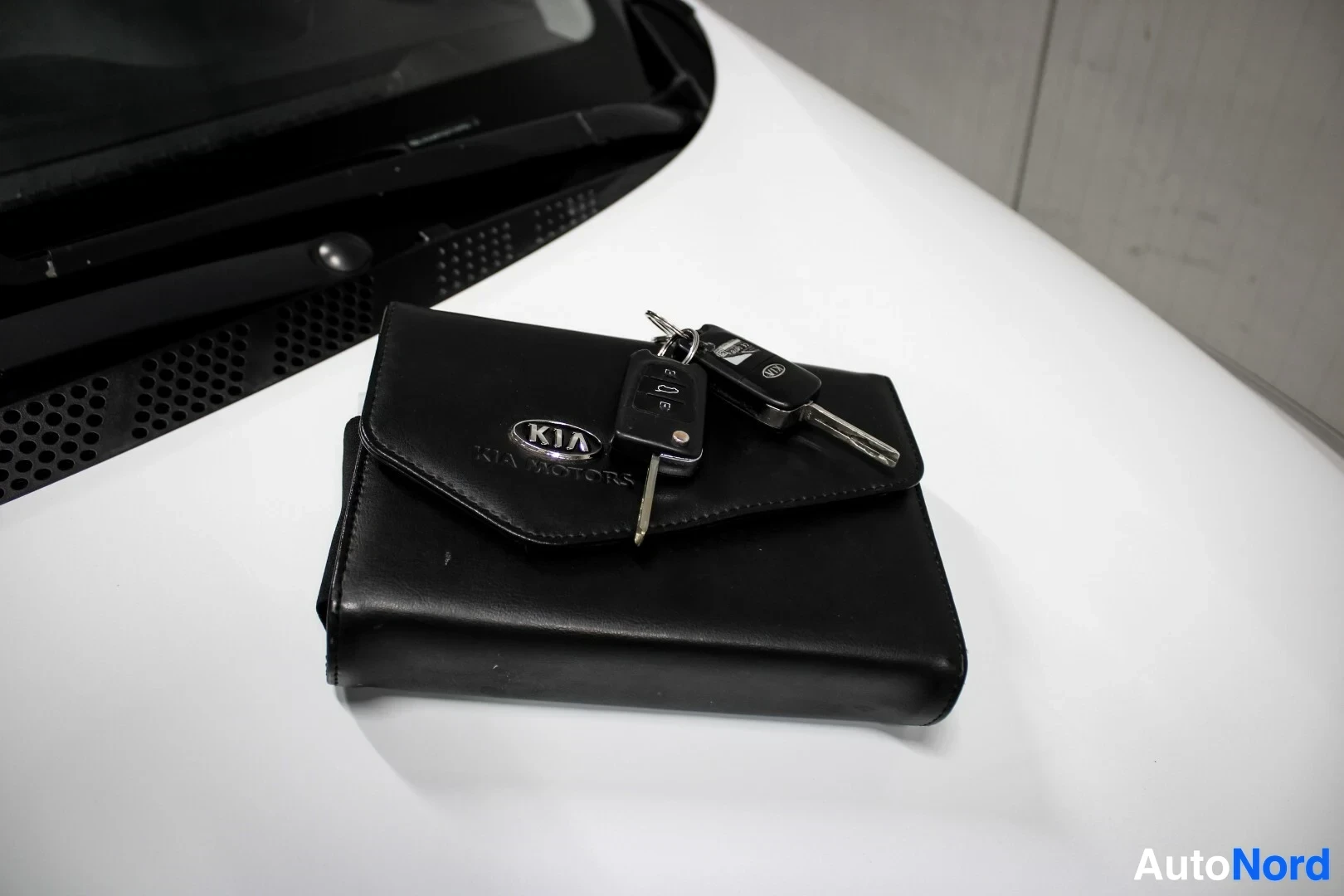 Yksityiskohta: Lähikuva kahdesta auton avaimesta, joissa toisessa on Kia-logo, ja mustasta Kia-kansiolaukusta auton konepellillä.
